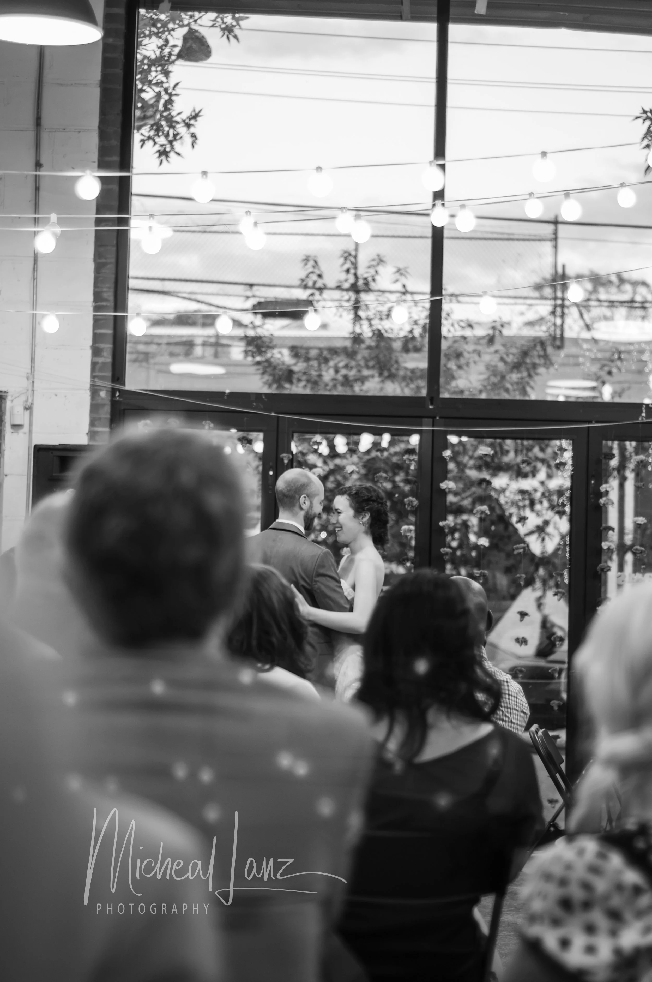 A wedding couple, a man and woman, sharing a moment while holding hands at their wedding reception inside a venue with large windows and string lights.