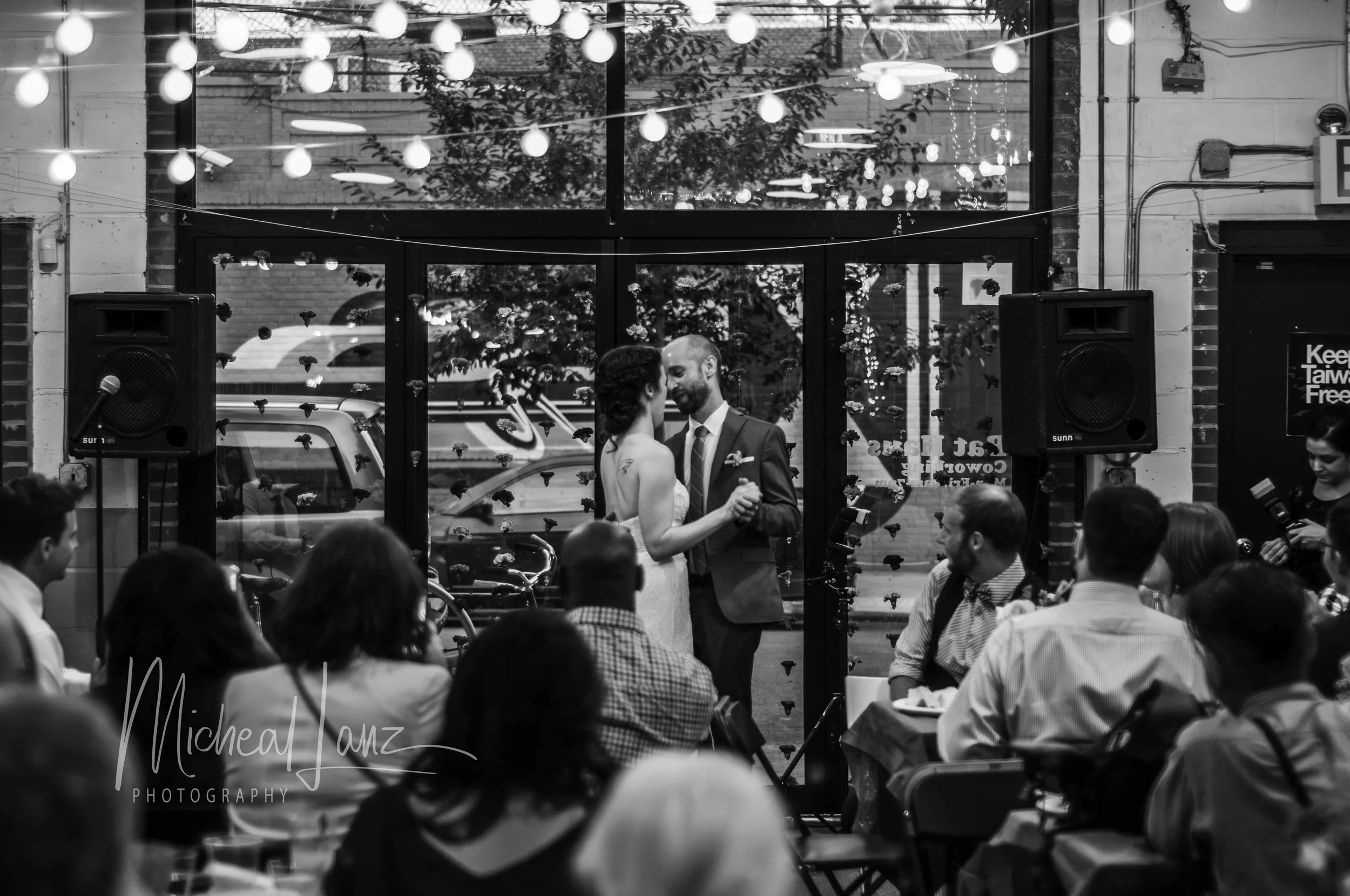 A black and white photo of a wedding reception with a couple dancing in front of large glass windows. The venue is decorated with string lights and floral arrangements. Guests are seated around tables, watching the couple.