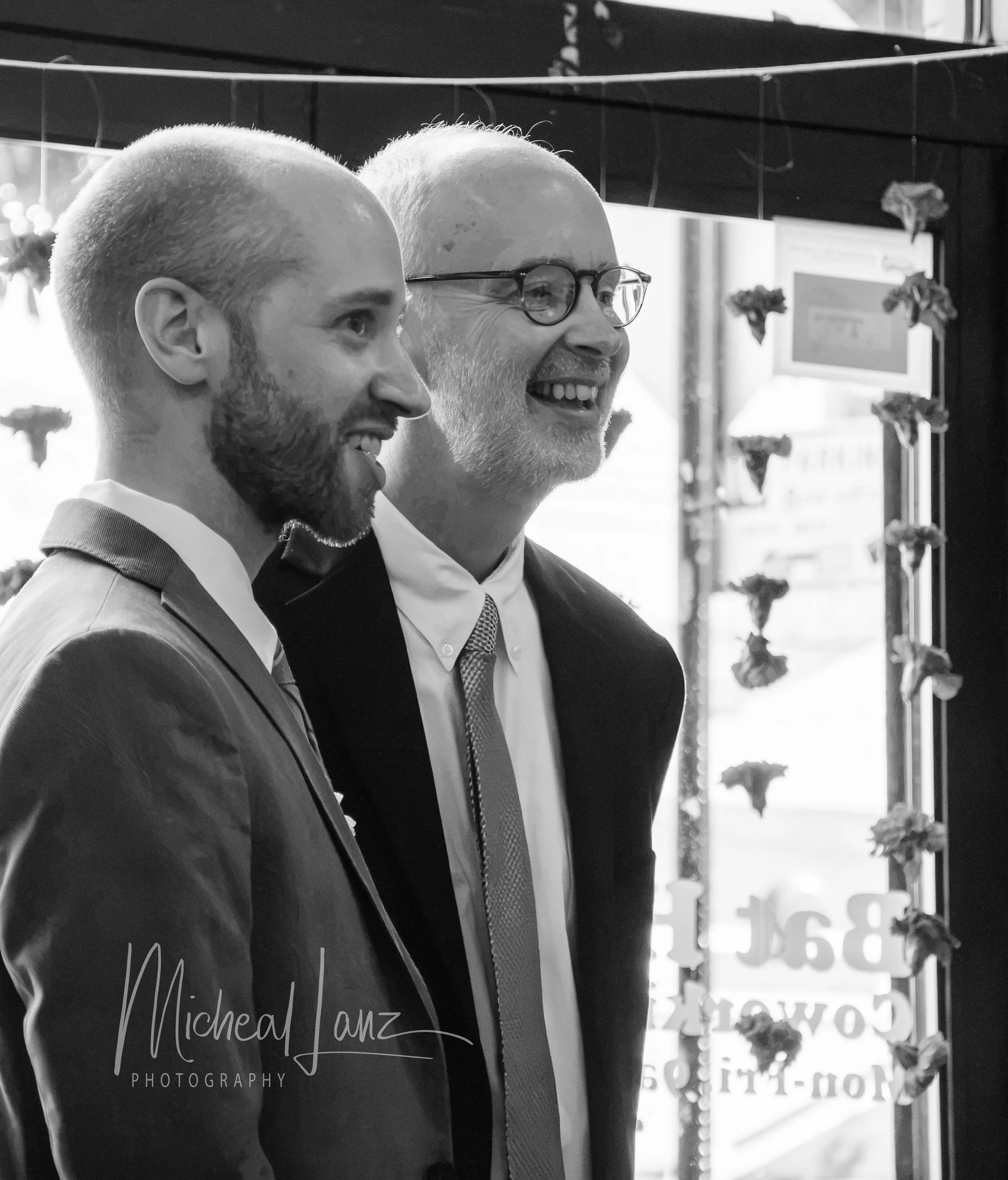 Father and son in suits smiling and standing side by side, with a storefront window and flowers in the background. On the sons wedding day.