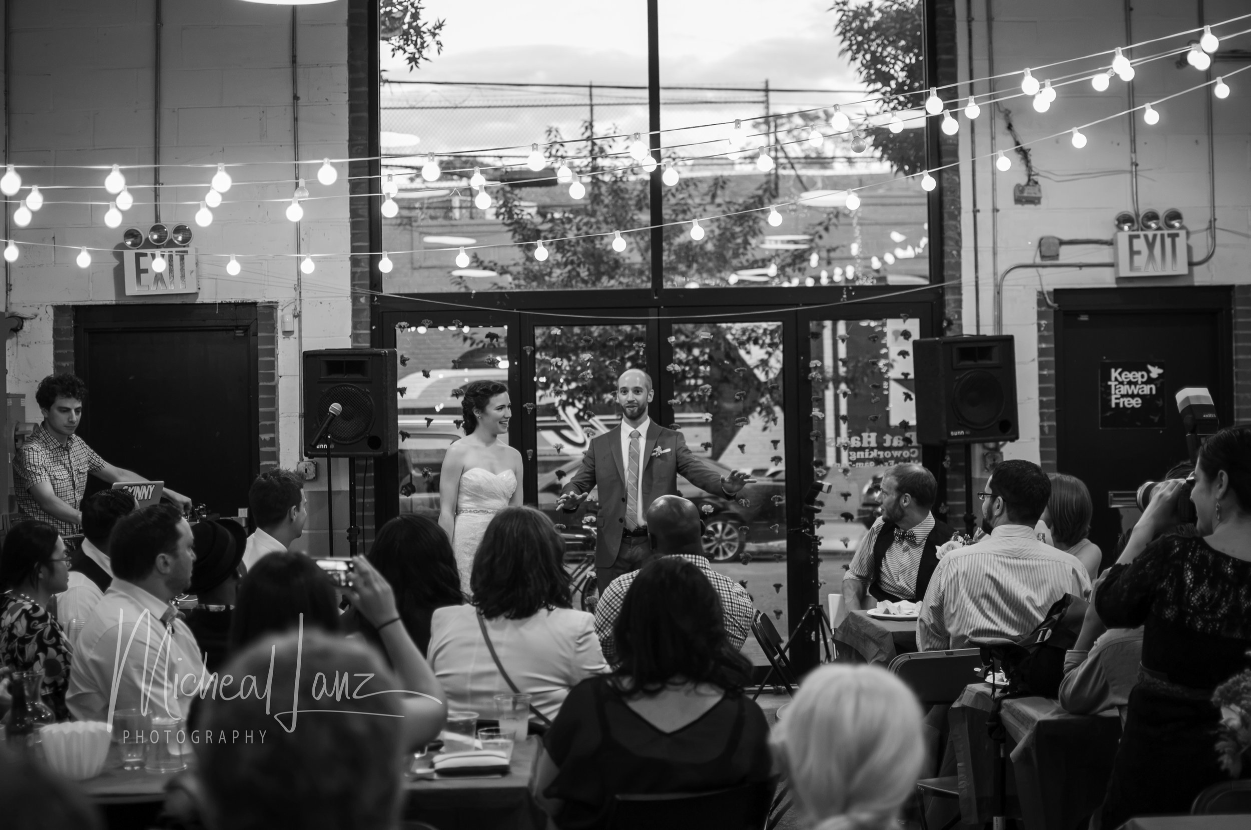 A wedding reception with a bride and groom standing side by side, surrounded by guests seated at tables. The venue has large windows, string lights, and an industrial style interior with exposed brick and concrete walls.