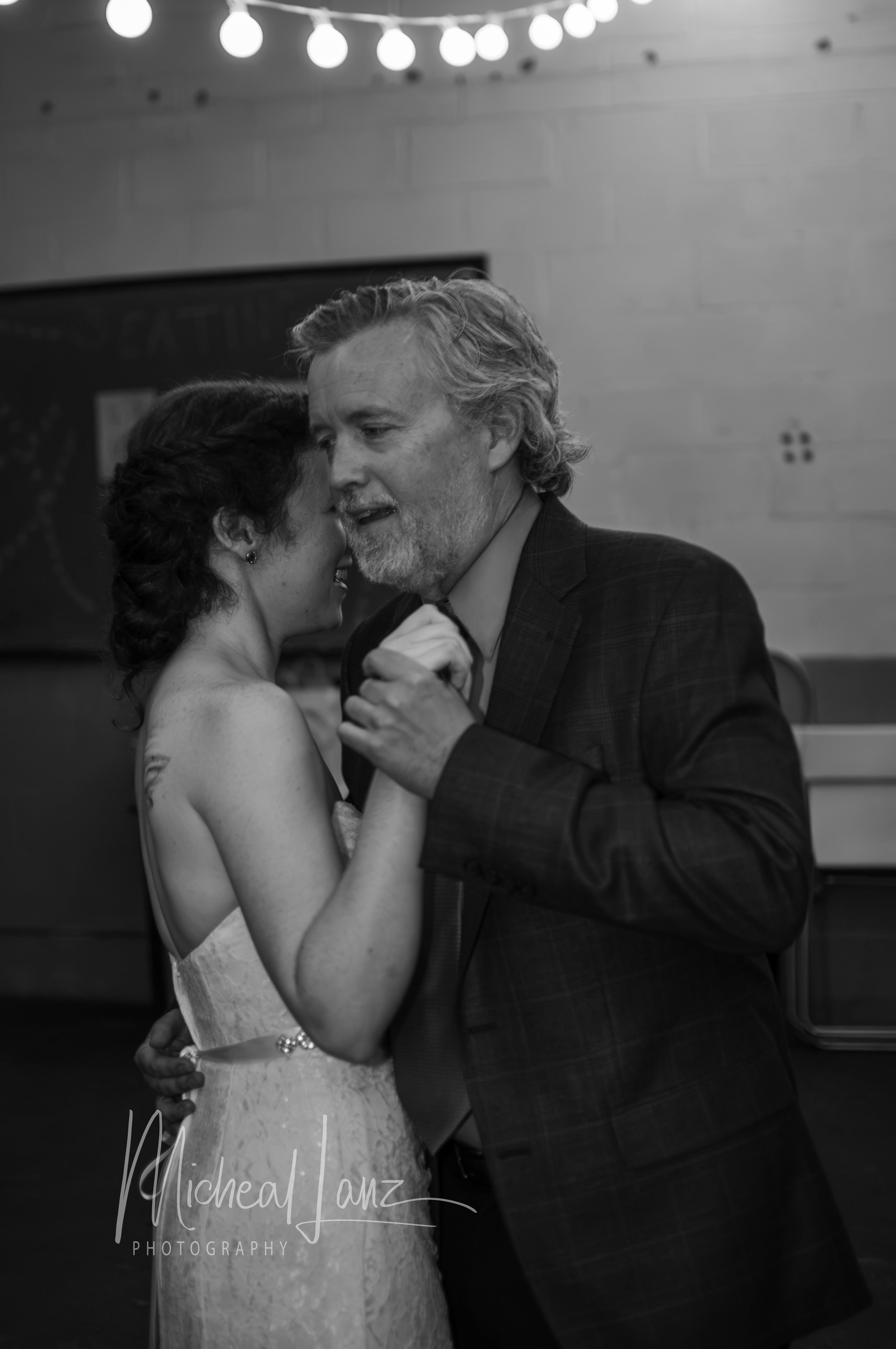 A black and white photo of a bride and her father sharing a dance at a wedding reception, dance floor in the background.