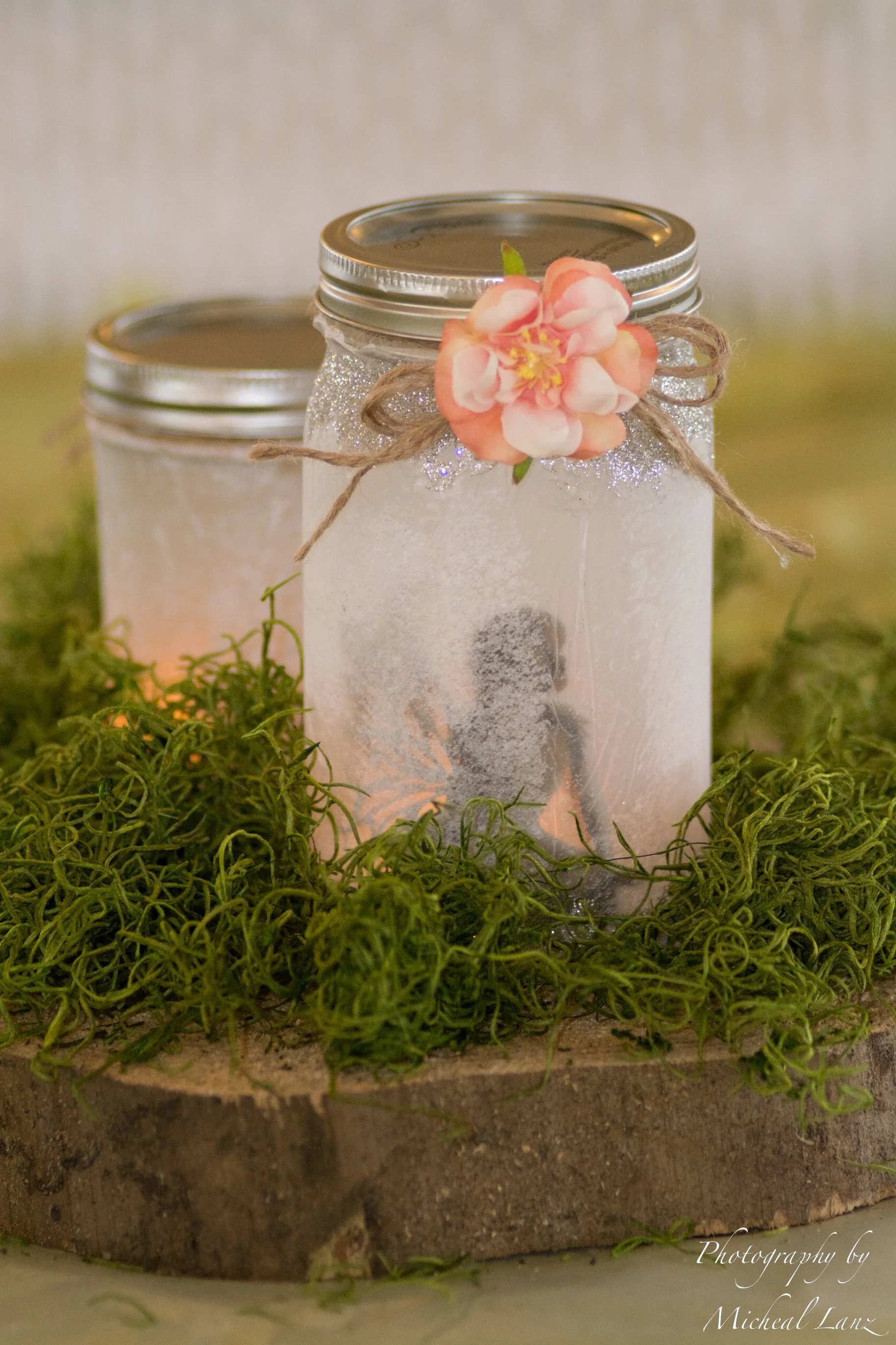 A photograph of two frosted glass jars with metal lids, wedding decorated with a pink flower and a twine bow, surrounded by green moss and sitting on a wooden surface.