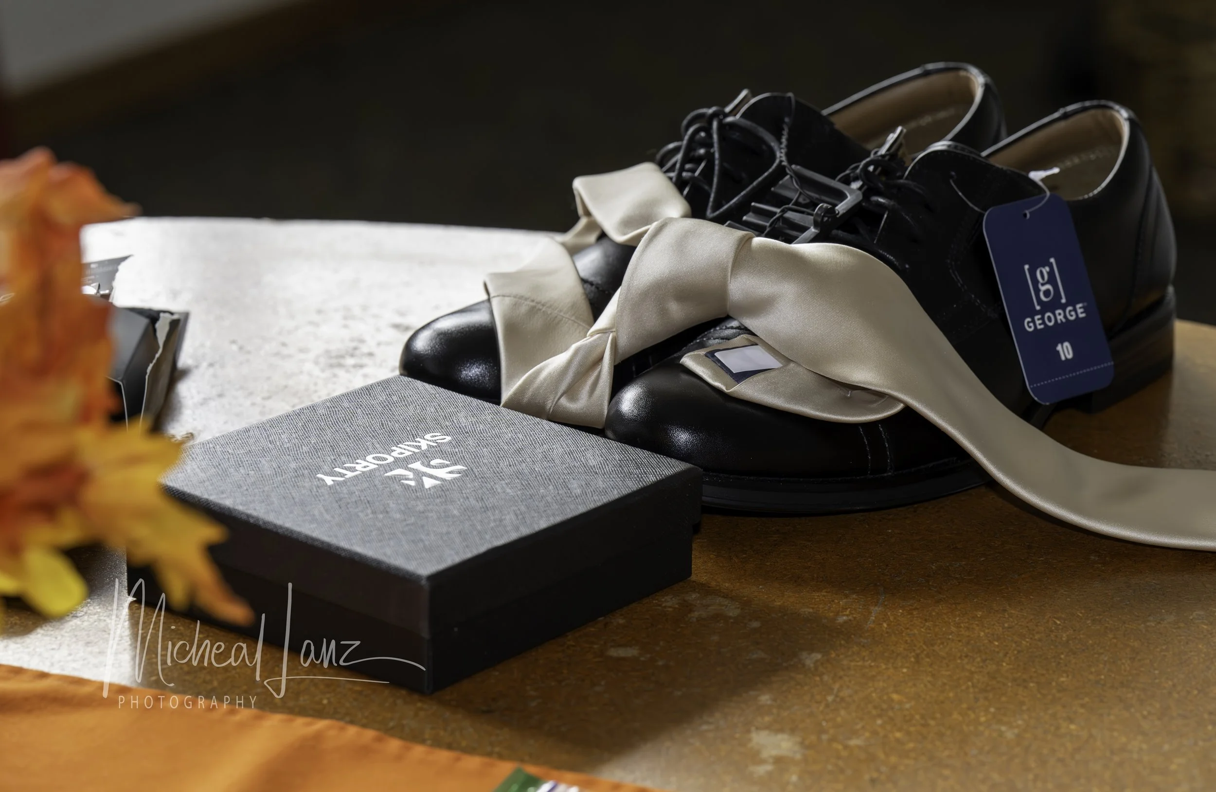 A pair of black leather shoes with satin ribbons tied in bows and a blue George size 10 tag, resting on a wooden surface alongside a closed gray book with white text and an orange flower on the left.