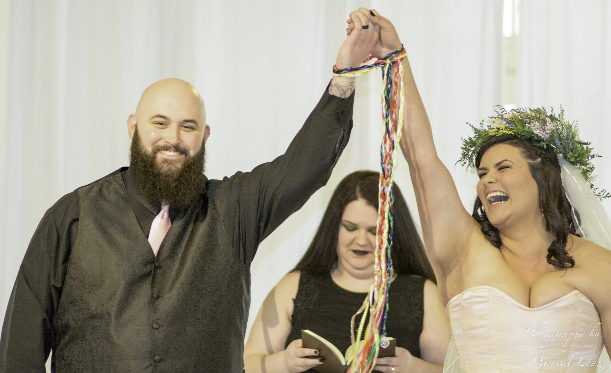 A wedding ceremony with a happy bride and groom holding hands and raising them together, smiling multiple women in the background, and floral decorations.