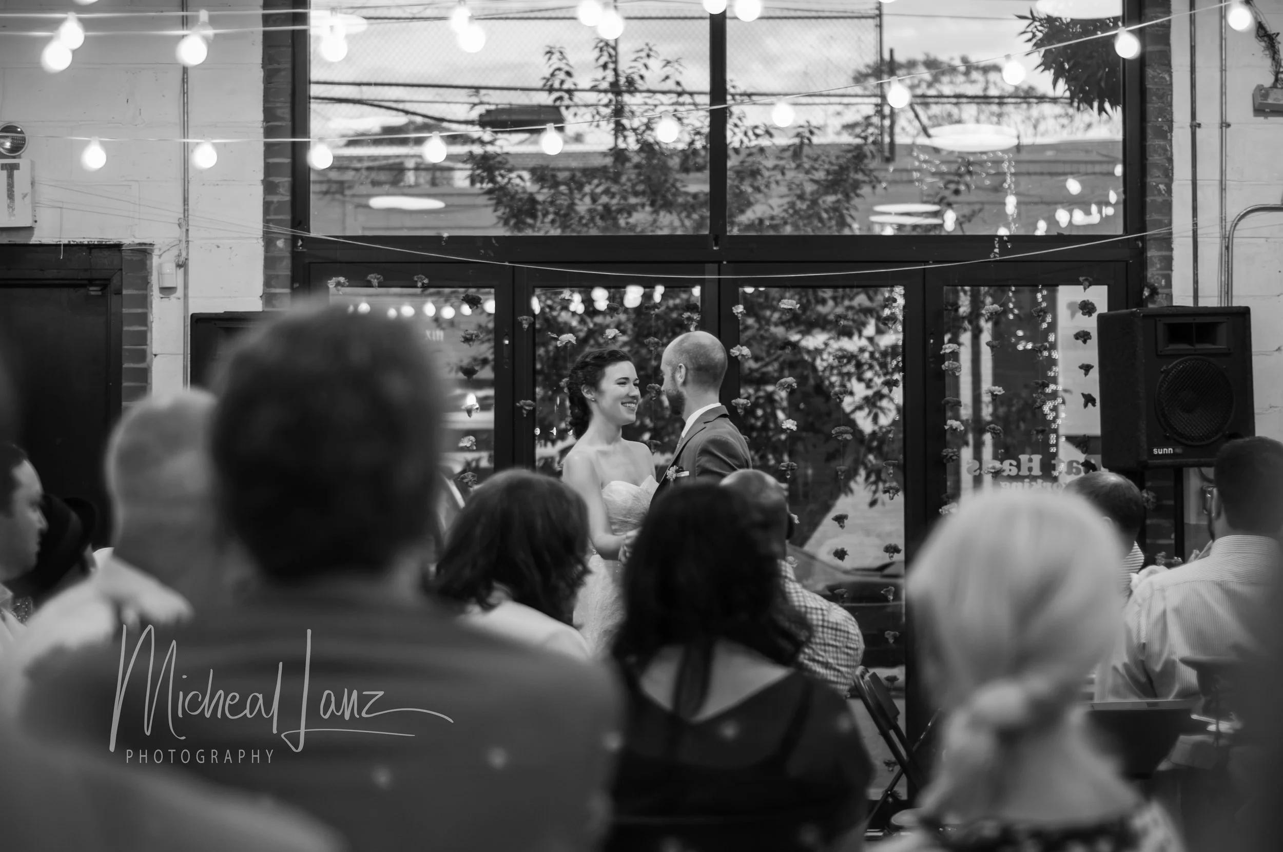 A black and white photograph of a wedding ceremony with a bride and groom standing together, smiling at each other inside a venue with string lights and large windows showing trees outside. The image is signed by Michael Lanz Photography.