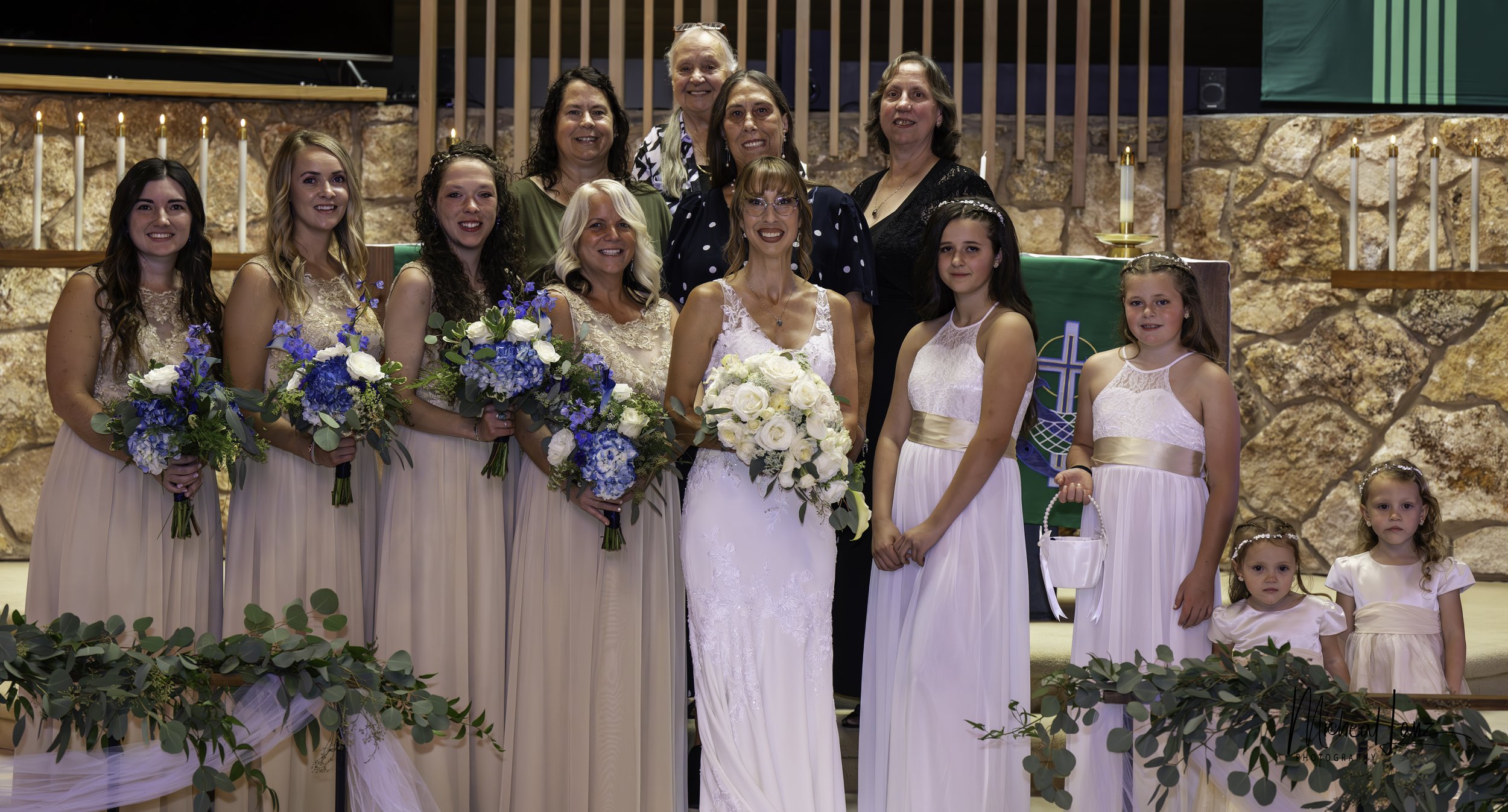 Group of women and girls at a wedding ceremony, with bridesmaids holding bouquets of blue and white flowers, standing in front of a stone wall with candles and green decor.