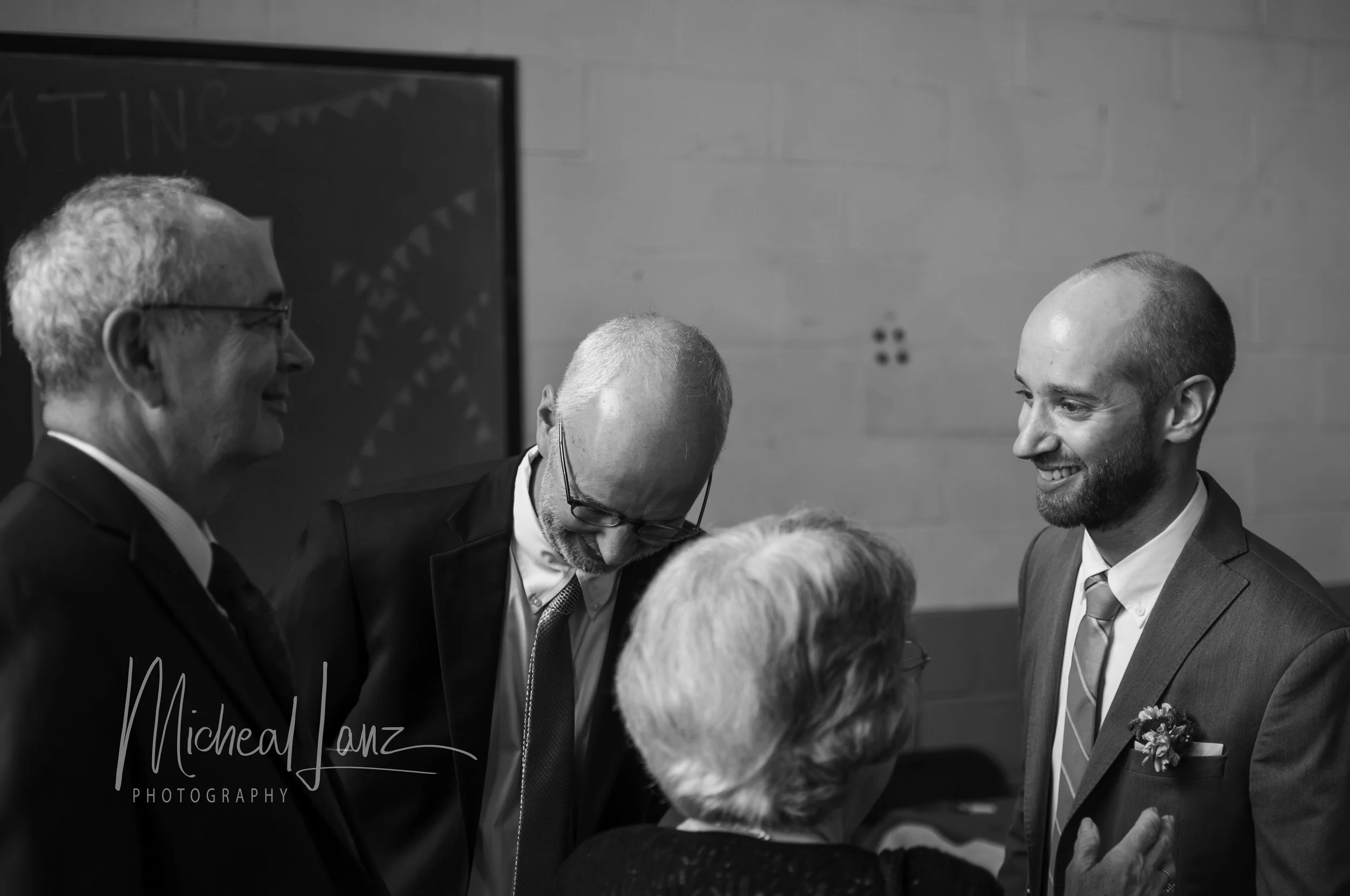 Black and white photo of men in suits at a formal event, smiling and talking to each other. One man is wearing glasses and has gray hair, another has a shaved head, and a third has a beard and is smiling. A woman with white hair is in the foreground.