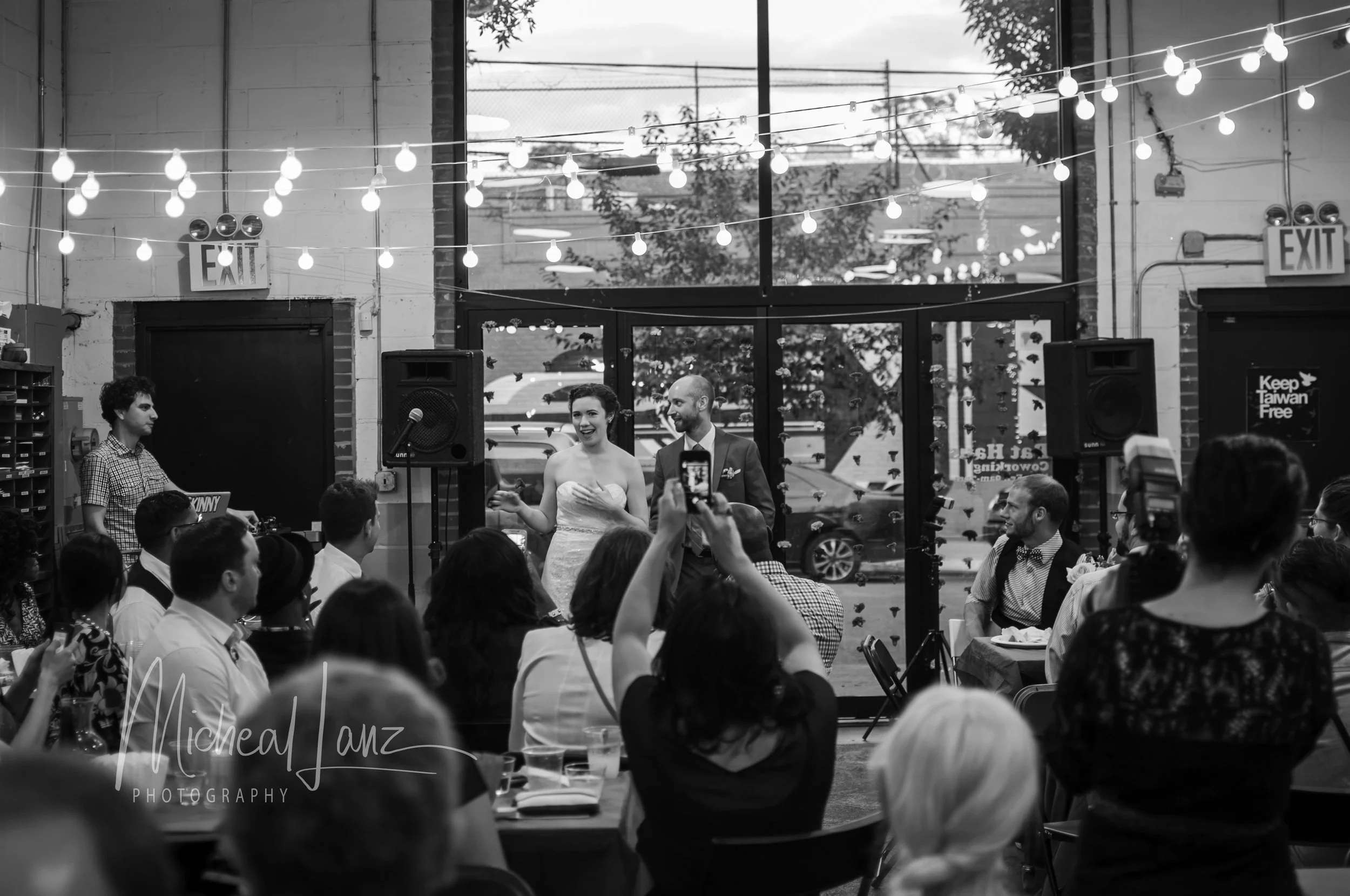 A wedding reception inside a venue with string lights hanging from the ceiling. The bride and groom are dancing in front of seated guests, with the bride smiling and the groom looking at her. There is a large window behind them showing outdoor trees 