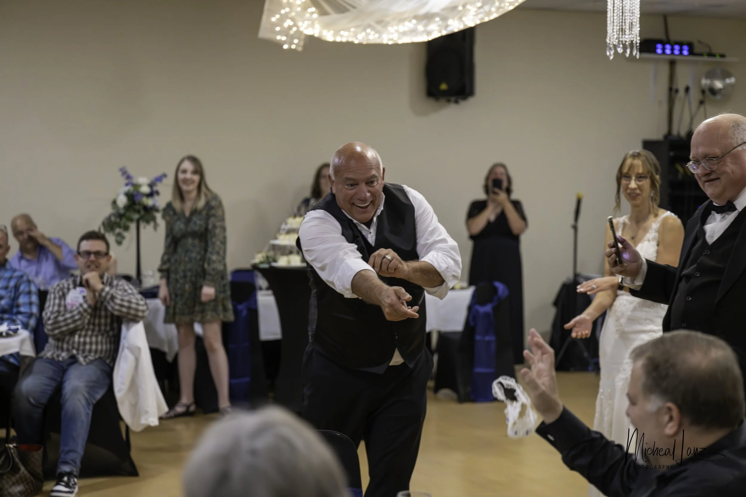 A man in a black vest and white shirt smiling and reaching out to a woman in a wedding dress at a wedding reception. Several guests are watching and smiling in the background.