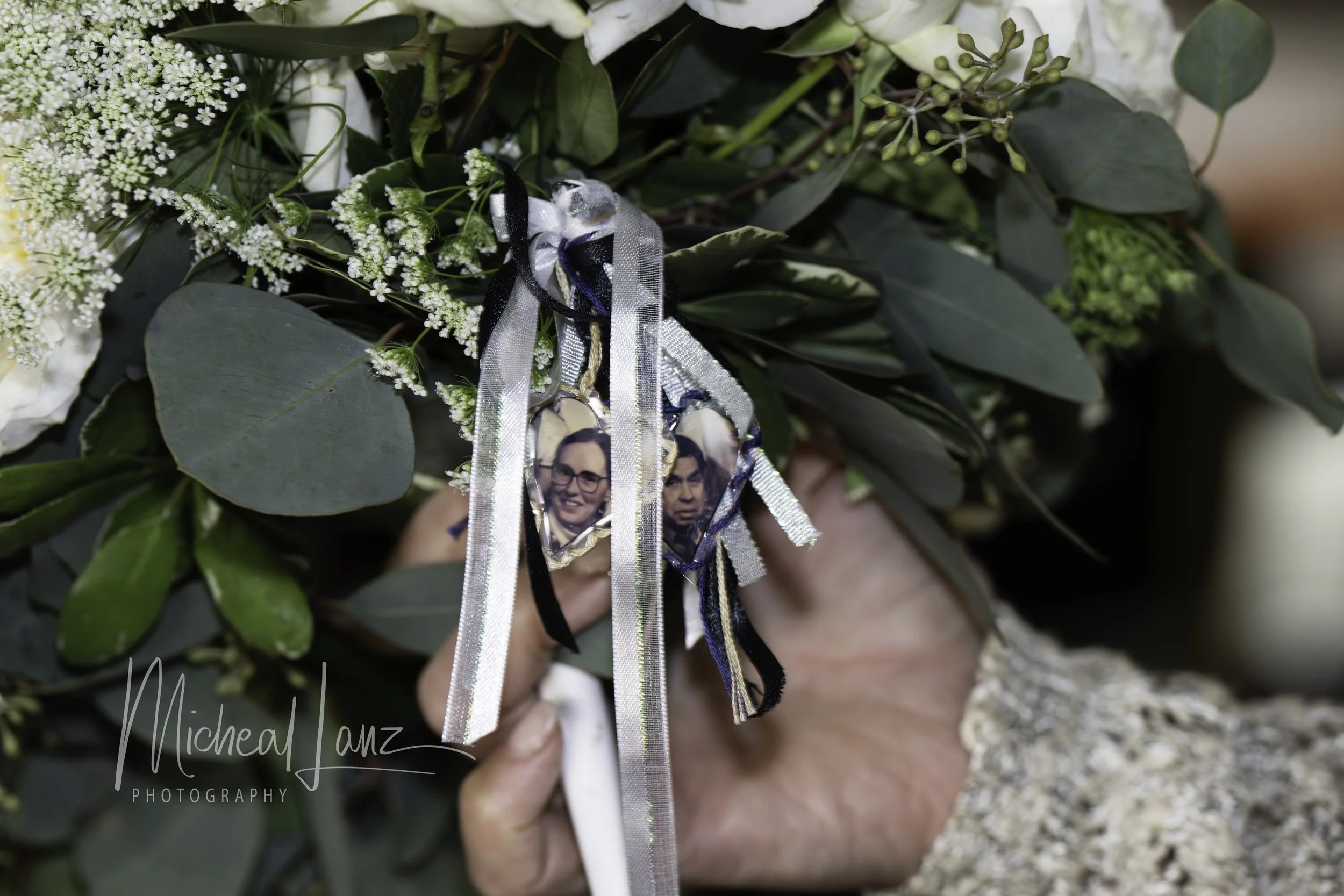 A close-up of a bouquet of white flowers and greenery held by a person. Attached to the bouquet are small photos of a woman and a man, along with ribbons in black, white, silver, and blue.
