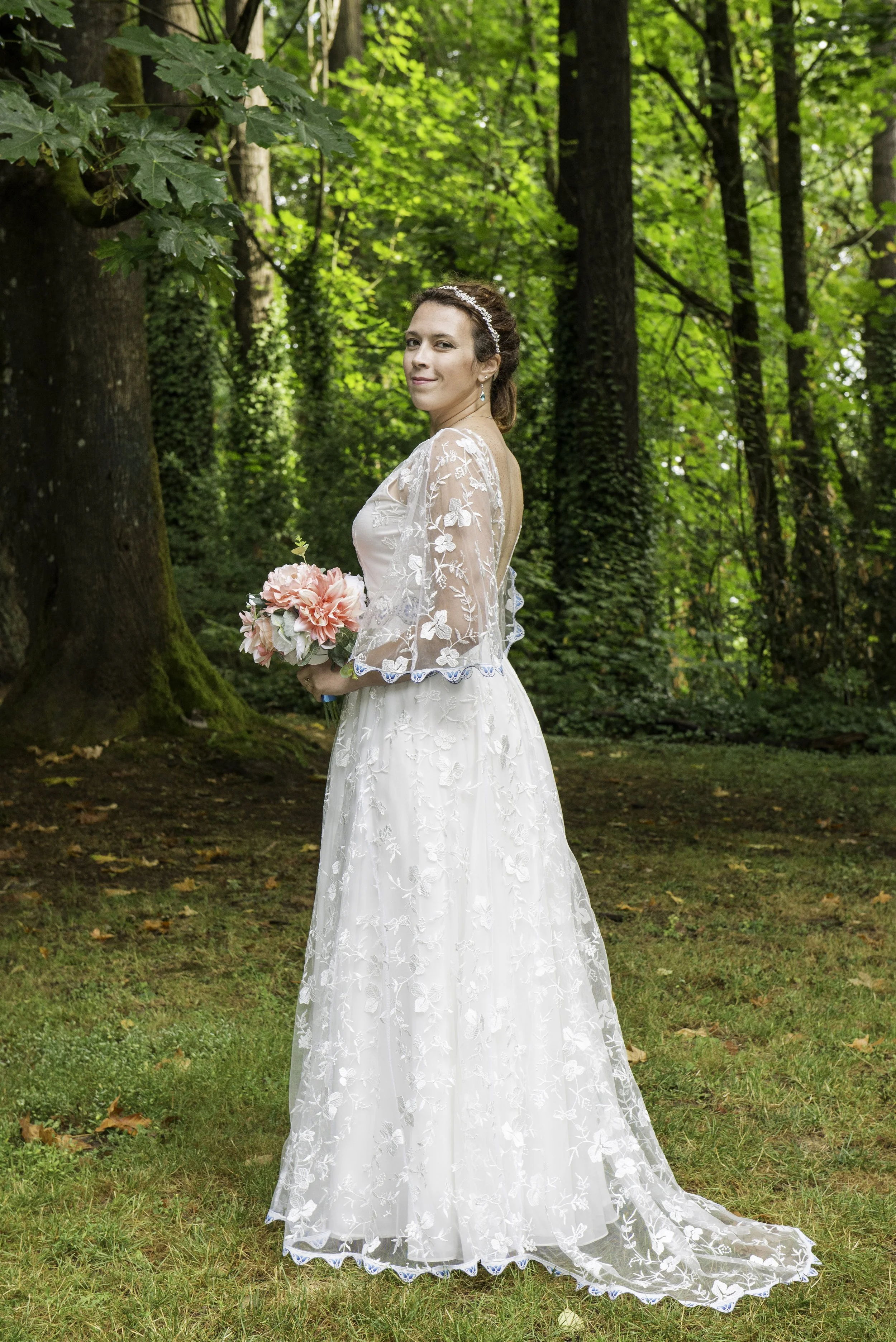 A woman dressed in a white lace wedding gown holding a pink and white bouquet standing outdoors in a green forest.