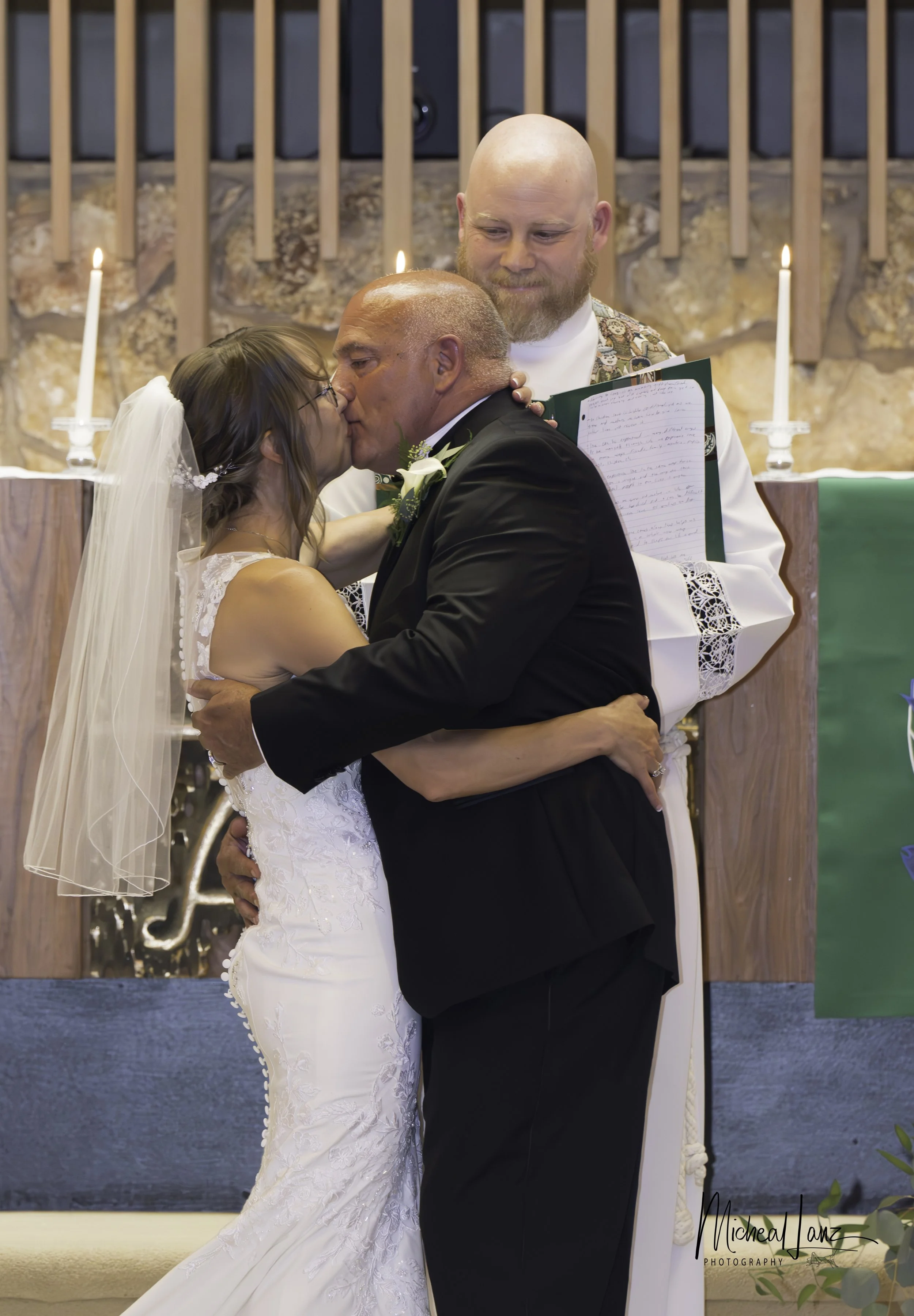 A bride and groom kiss during their wedding ceremony, with an officiant standing behind them holding a folder of papers. The bride wears a white lace wedding dress and veil, and the groom is in a black suit with a white boutonniere. Candles and a sto