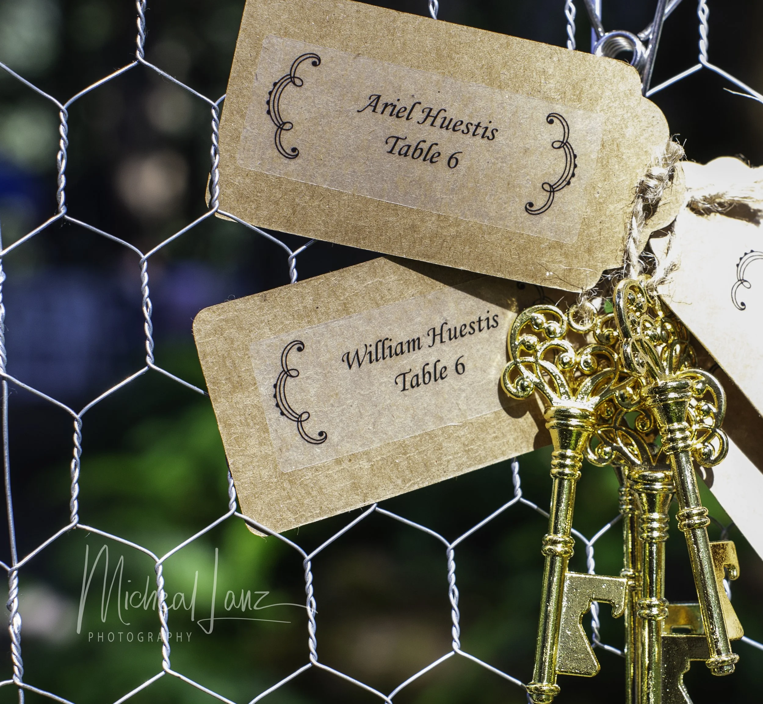 A close-up photo of a metal wire fence with a brown paper reservation sign that reads 'Ariel Huestis, Table 6'. A small section of a decorative, gold-colored key with a fancy bow knot is hanging on the fence next to the sign.
