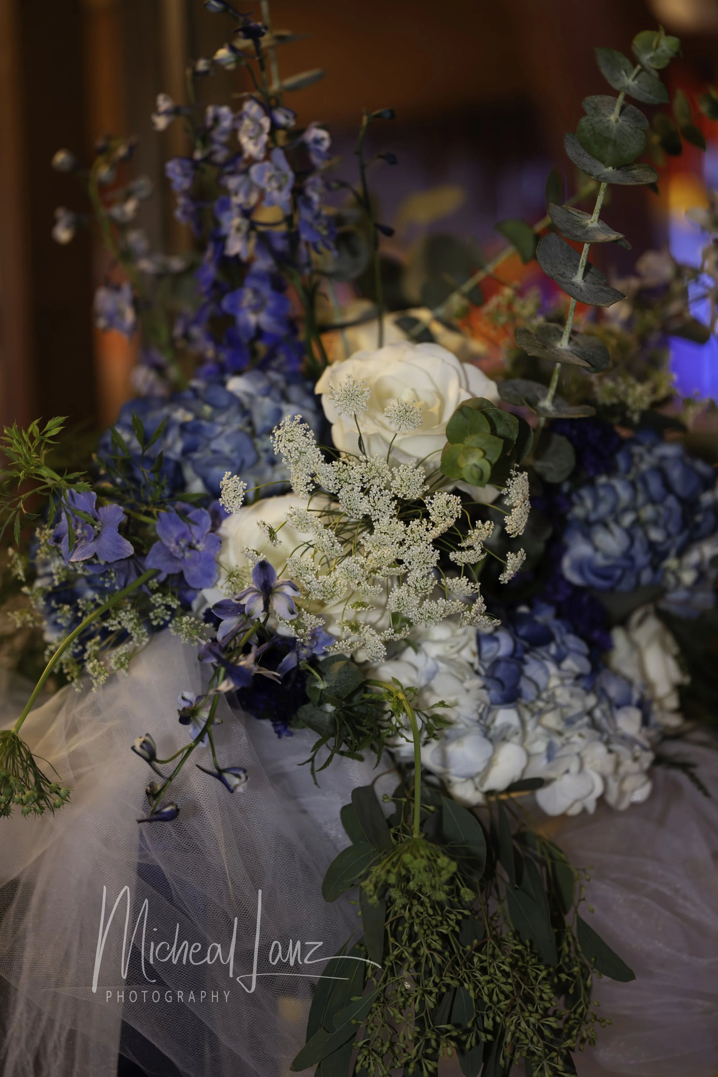 A floral bouquet with white roses, blue hydrangeas, delicate white flowers, and green foliage, wrapped in white tulle. 