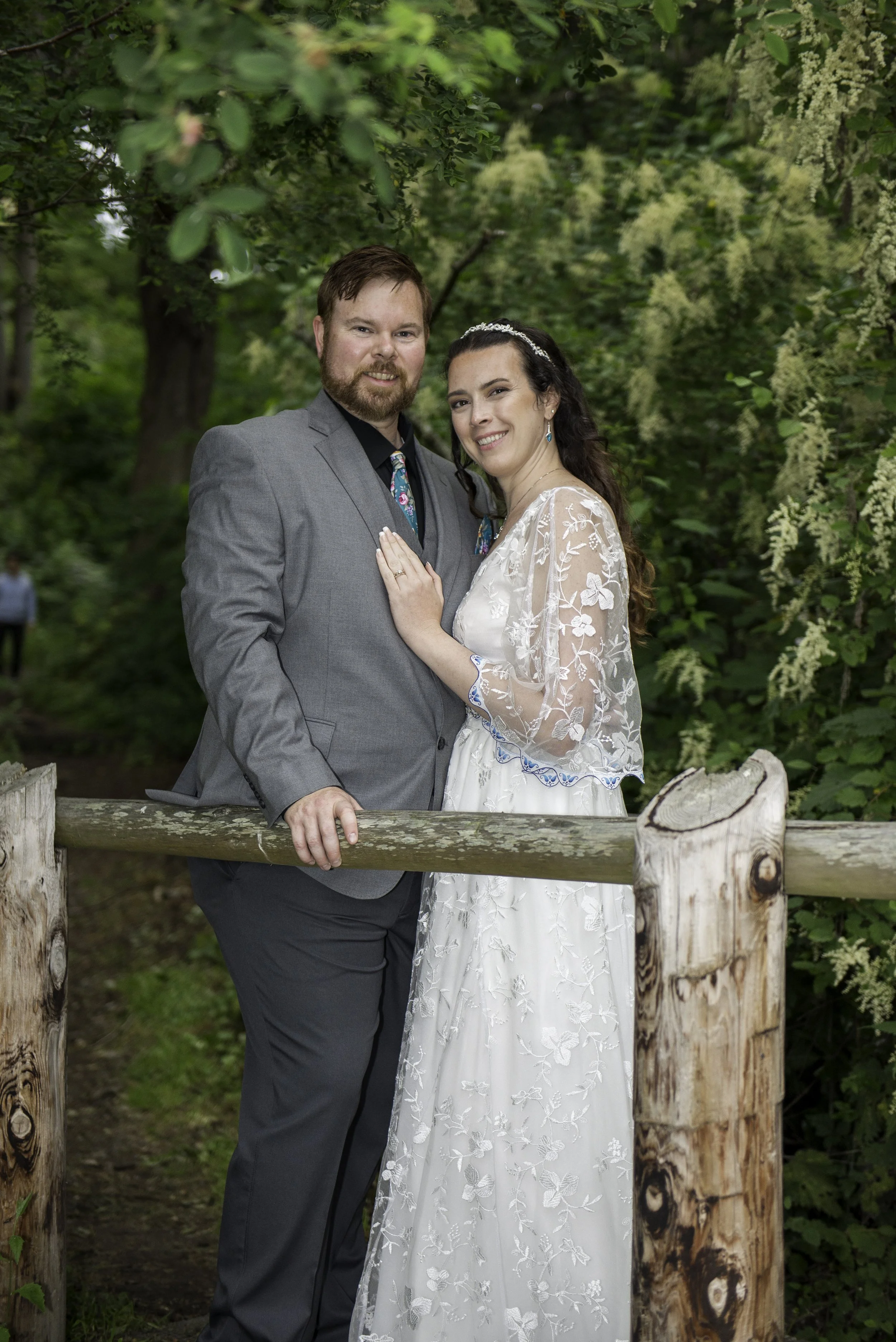 A newlywed couple posing outdoors in a green park, standing behind a wooden railing with lush trees in the background. The groom wears a gray suit with a colorful tie, and the bride wears a white lace wedding gown with floral embroidery. The bride ha