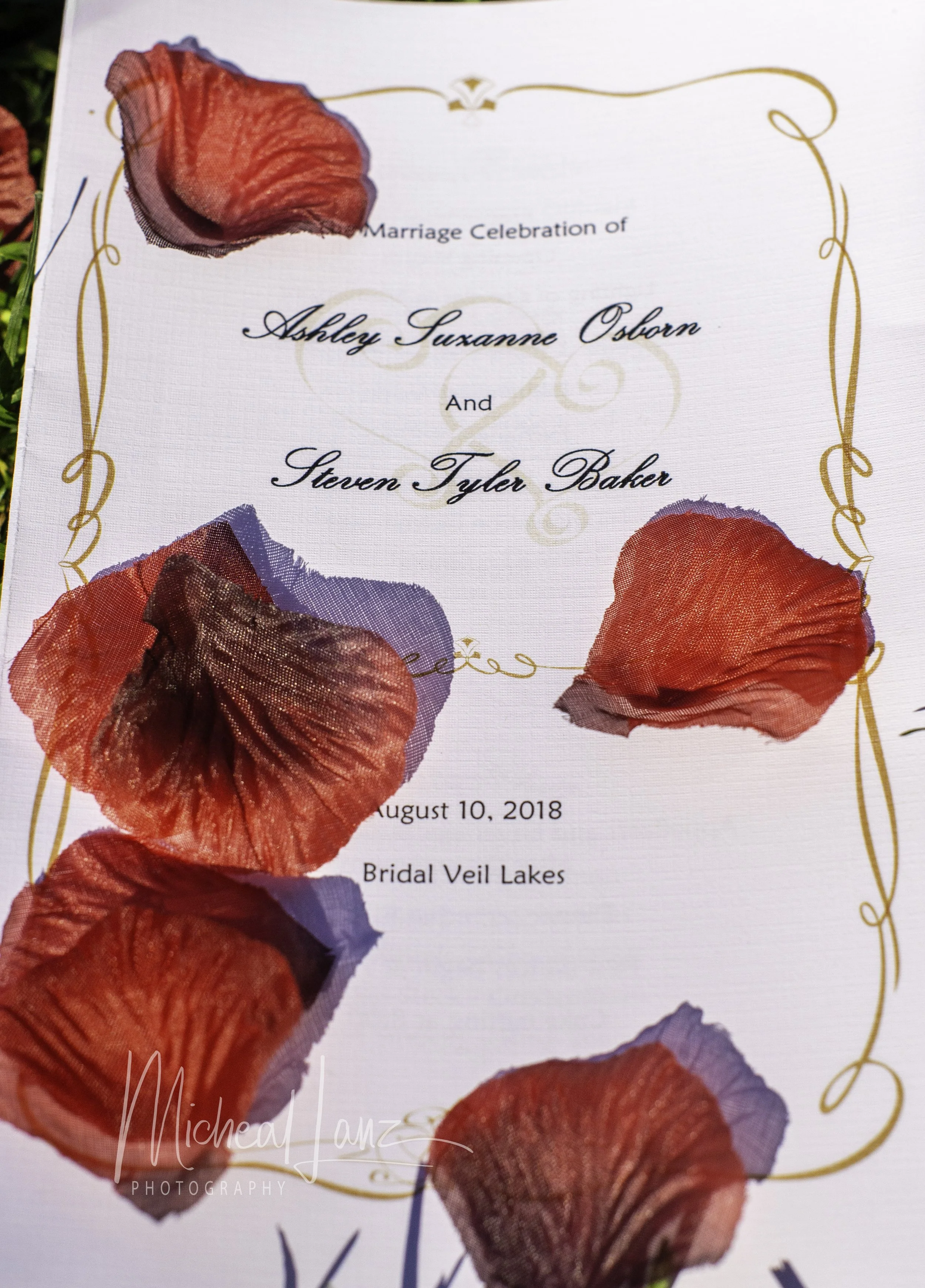 A wedding invitation with red flower petal decorations on a white background, including details of the marriage celebration of Ashley Suzanne Osborn and Steven Tyler Baker on August 10, 2018, at Bridal Veil Lakes.