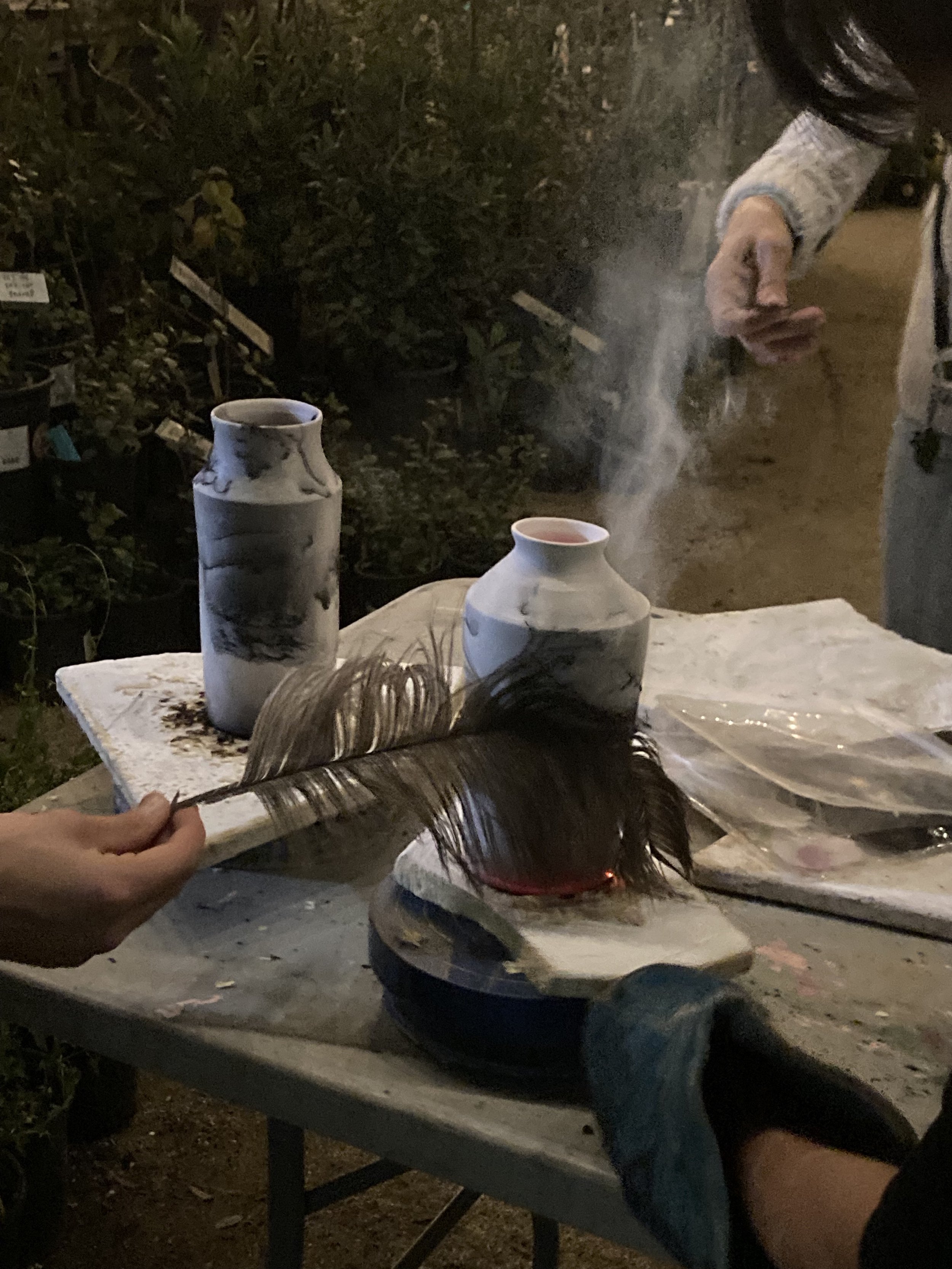 People handling a bird with long, dark feathers on a table near two ceramic vases at night.