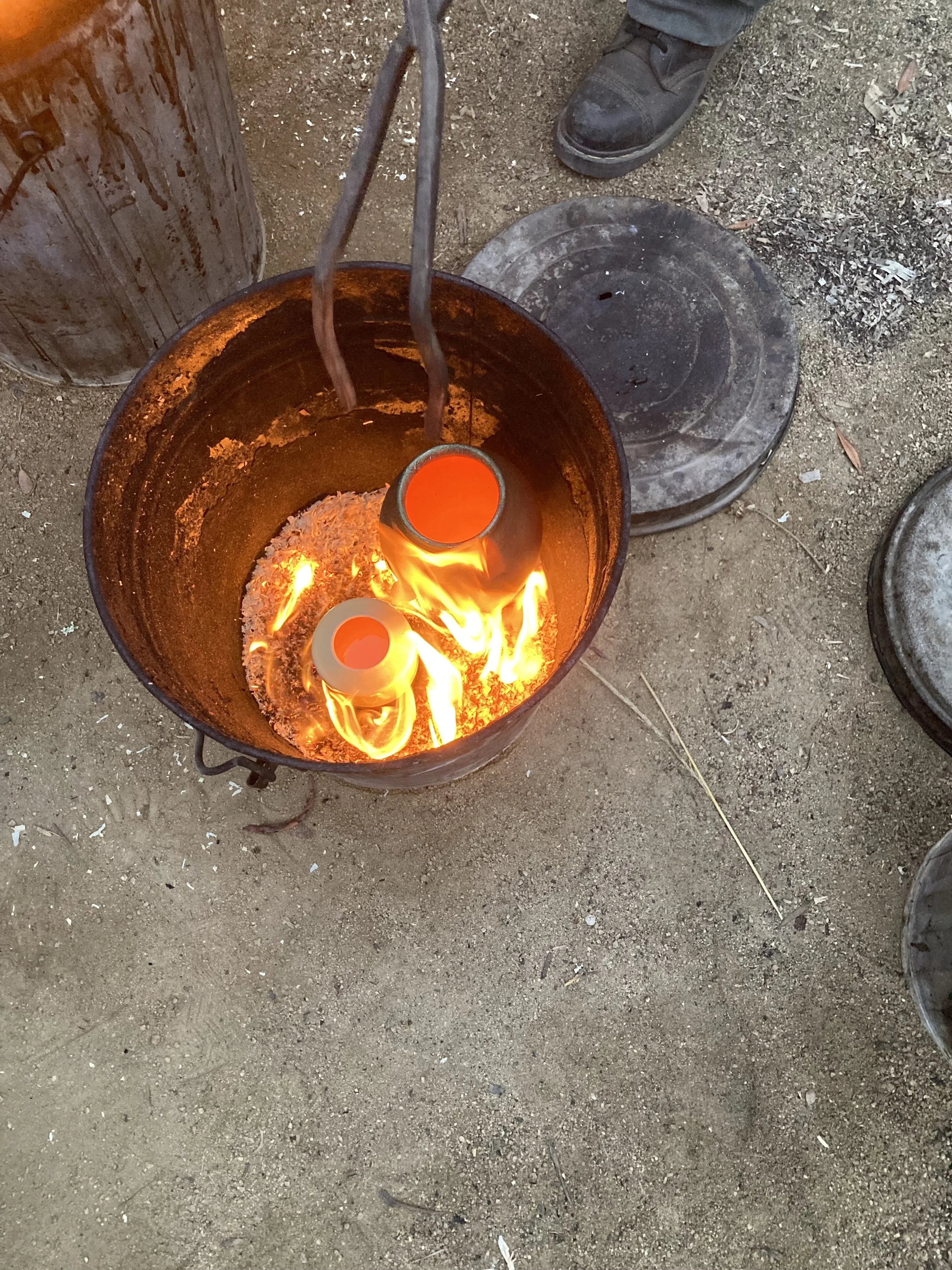 Two small red ceramic vessels being heated in a black metal barrel over an open flame, outdoors with dirt ground and covered by several black metal plates.