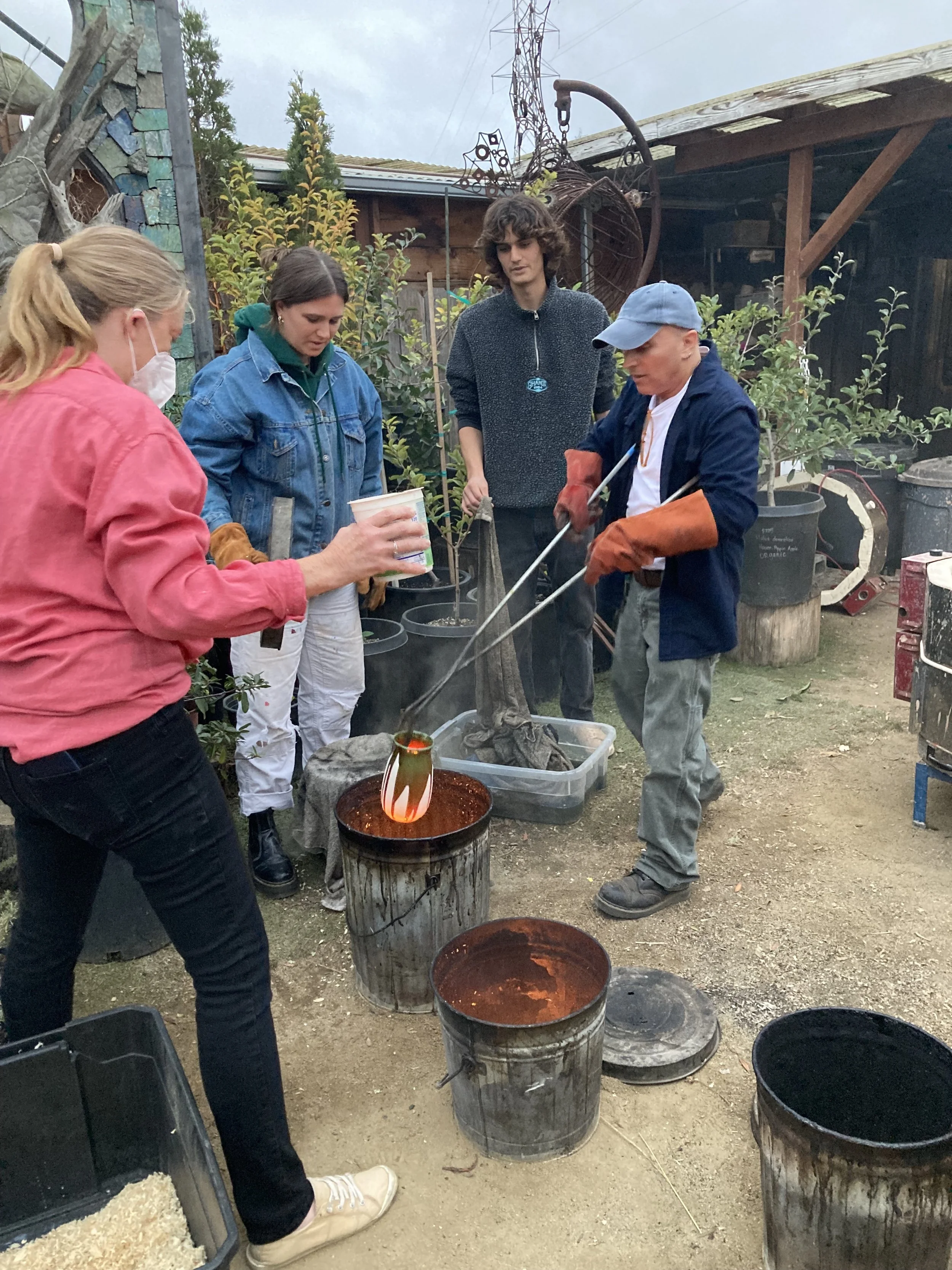 Group of five people outdoors, some wearing gloves and masks, handling a glass art piece over a forge with buckets nearby, with trees and a wooden shed in the background.