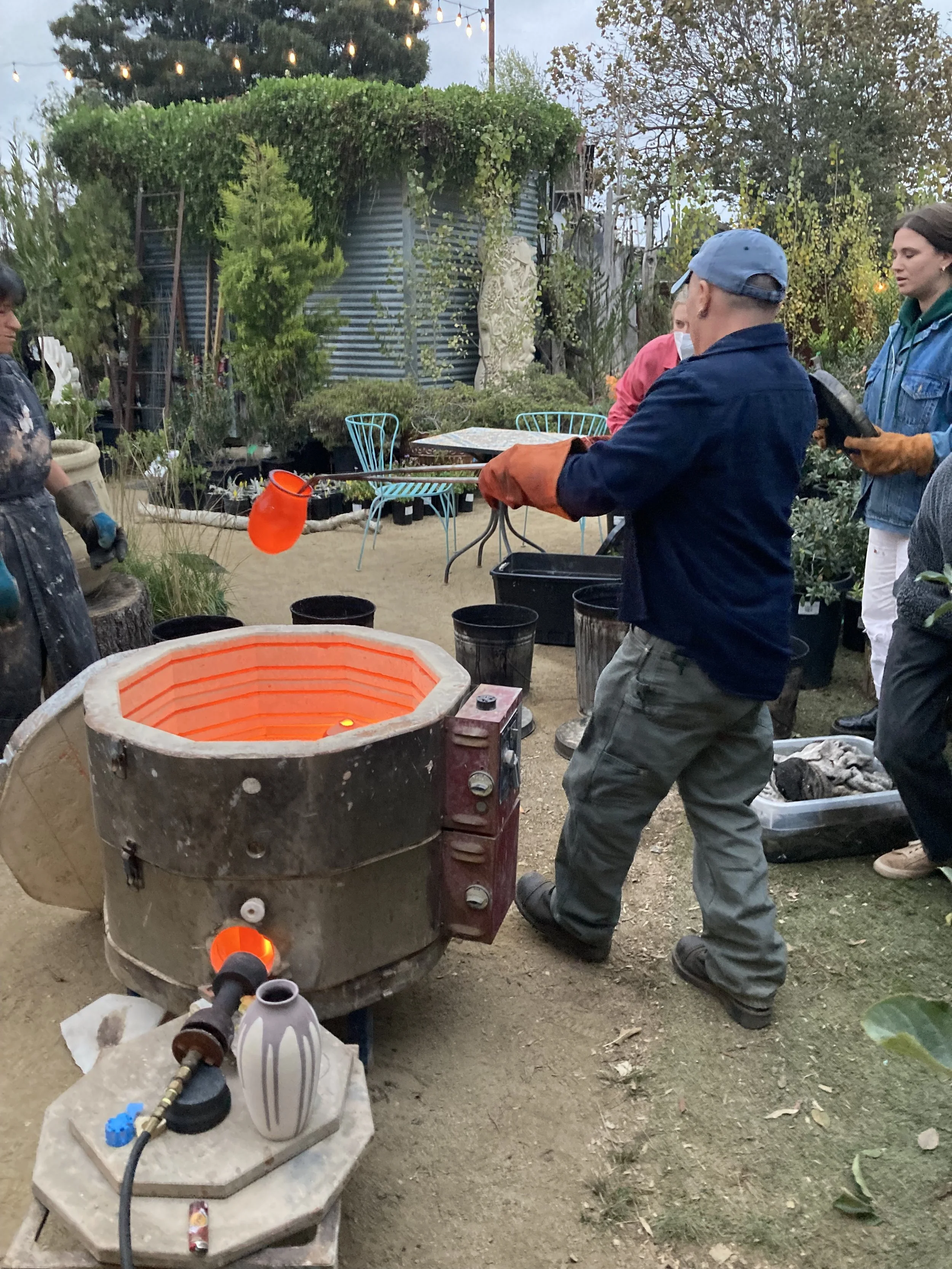 People working in a pottery studio with a kiln on the ground, a person handling clay or tools, and various pots and supplies around, set outdoors in a garden area with trees and string lights.