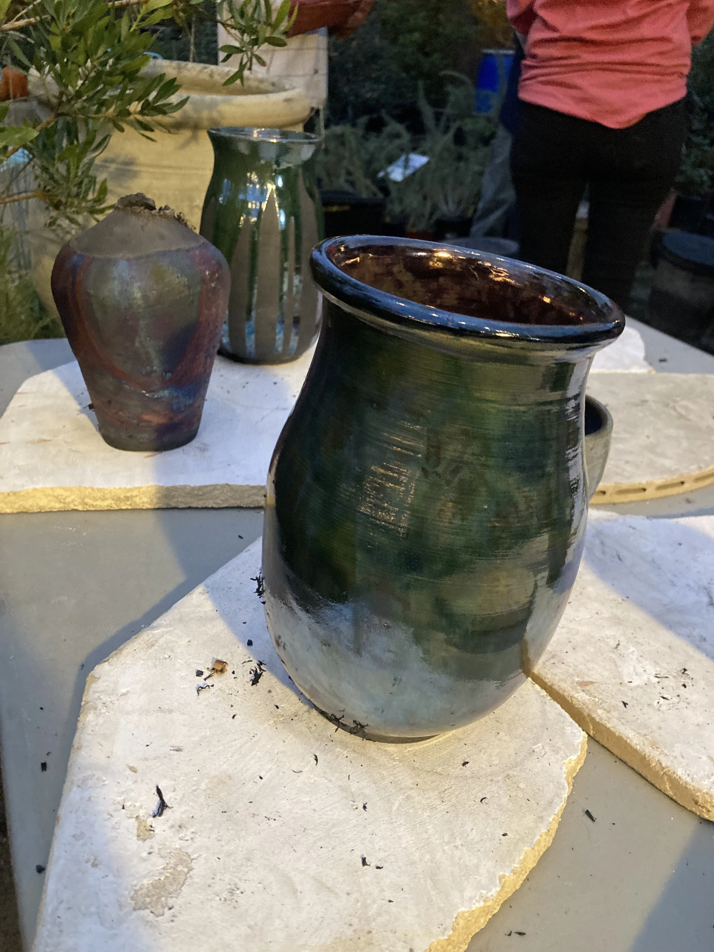 Colorful glazed ceramic vases on a stone surface outdoors, with greenery and a person in a red shirt and black pants in the background.