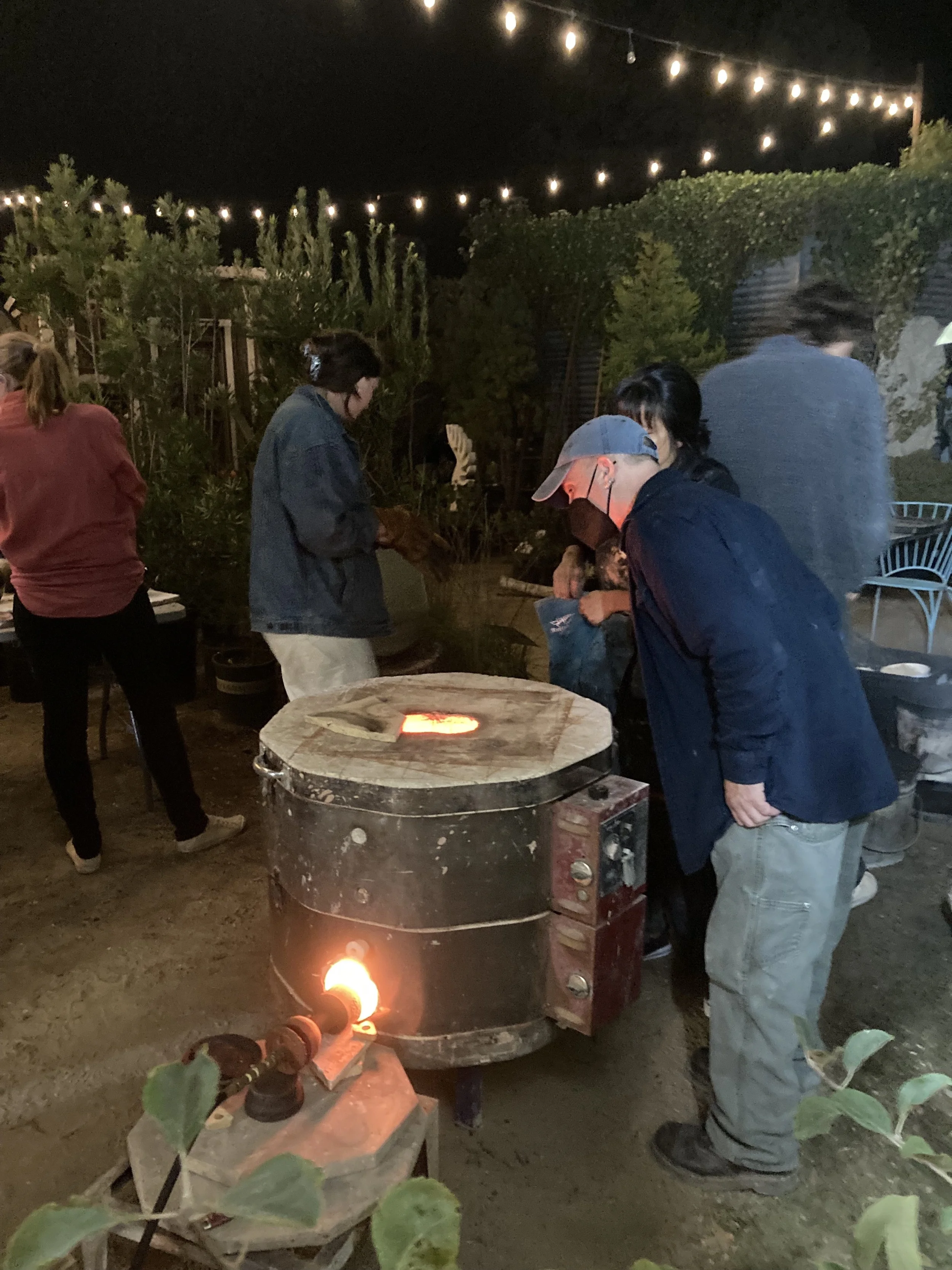 People working at a glassblowing studio at night, with string lights overhead and a small forge in front of them.