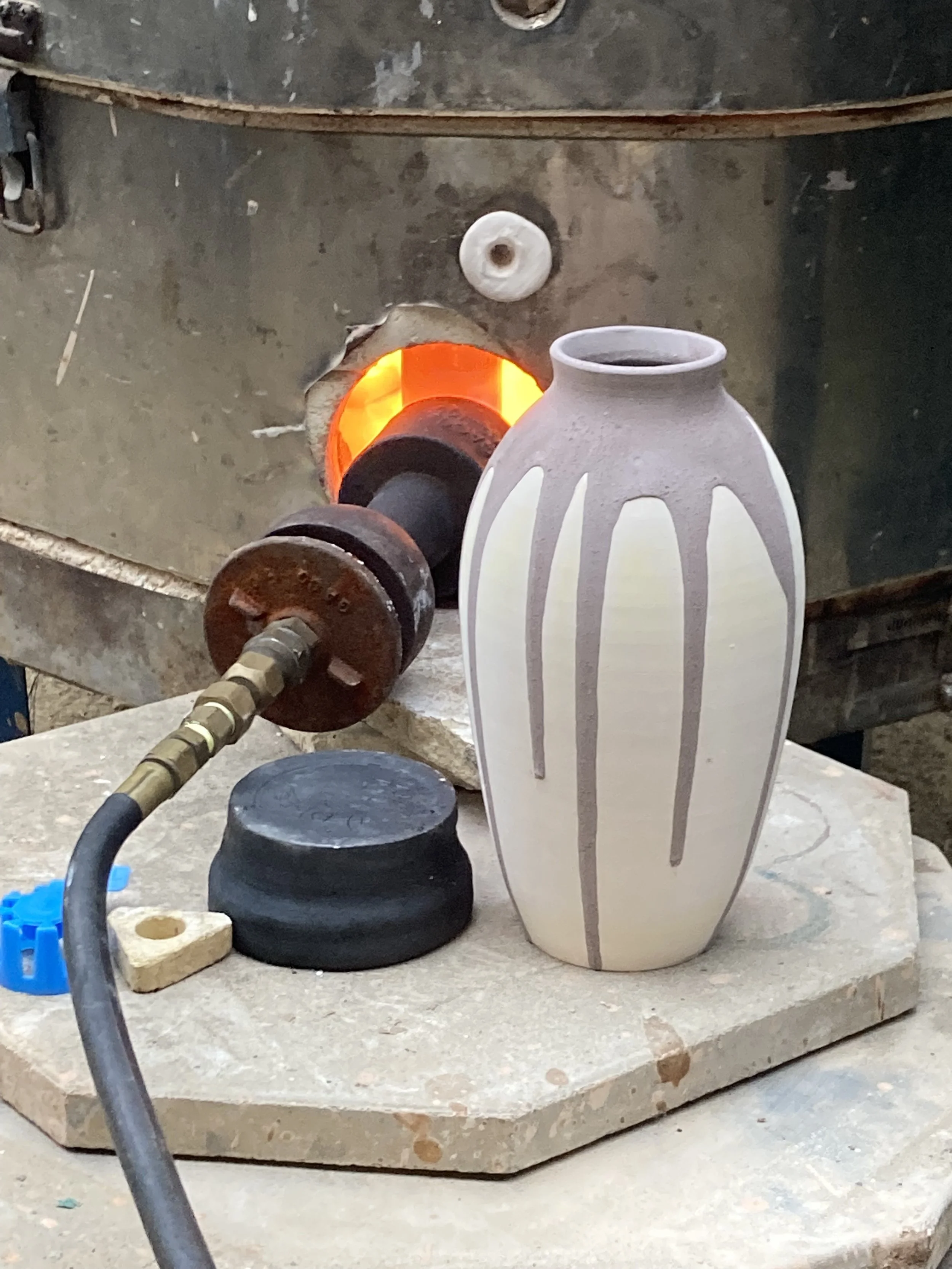 Ceramic vase on a pottery wheel in a kiln with glowing orange interior, surrounded by pottery tools and a black stand.