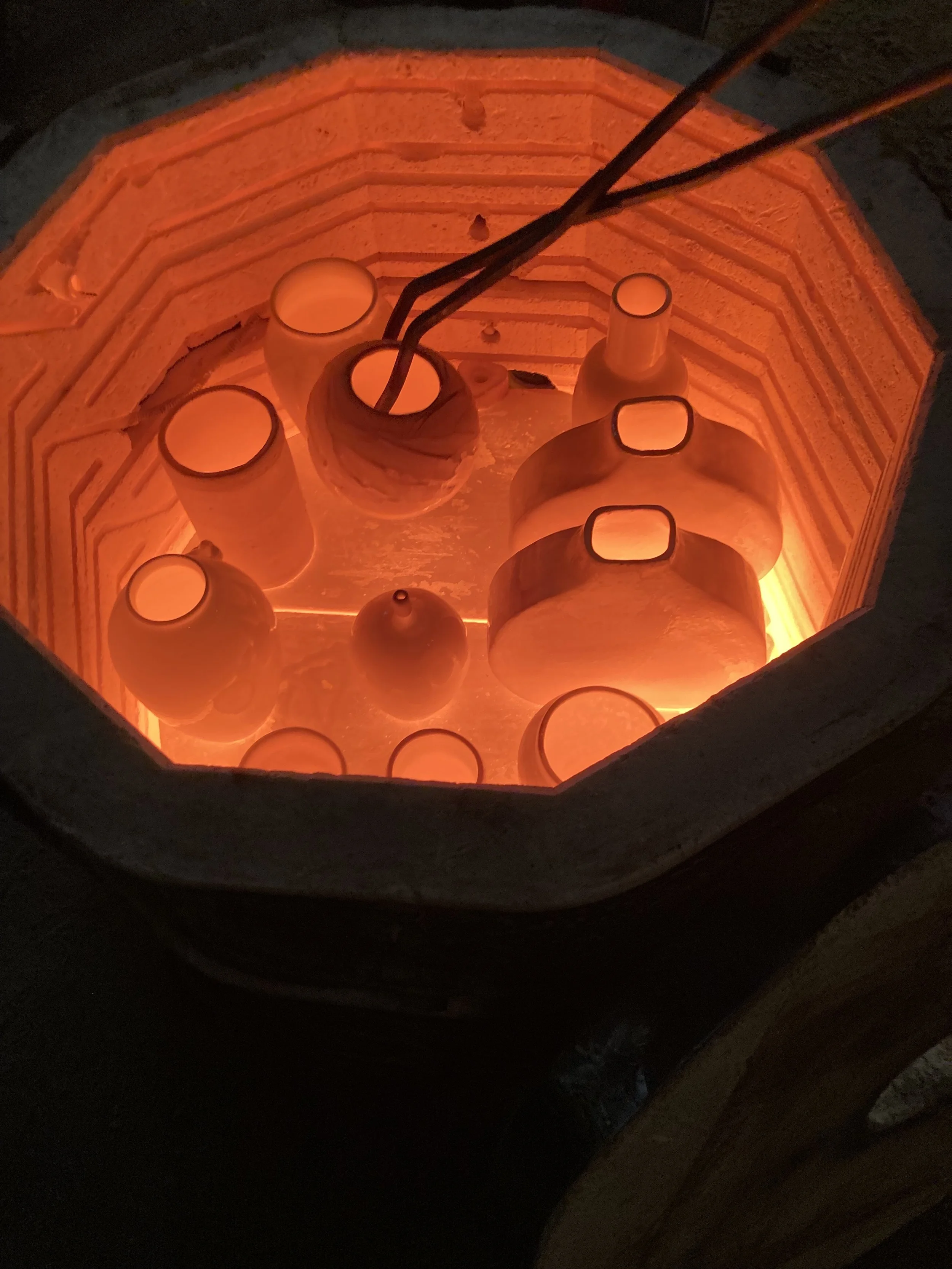 Lit kiln with orange glowing embers, containing several ceramic vases and bowls, some with open tops and some with narrow necks.