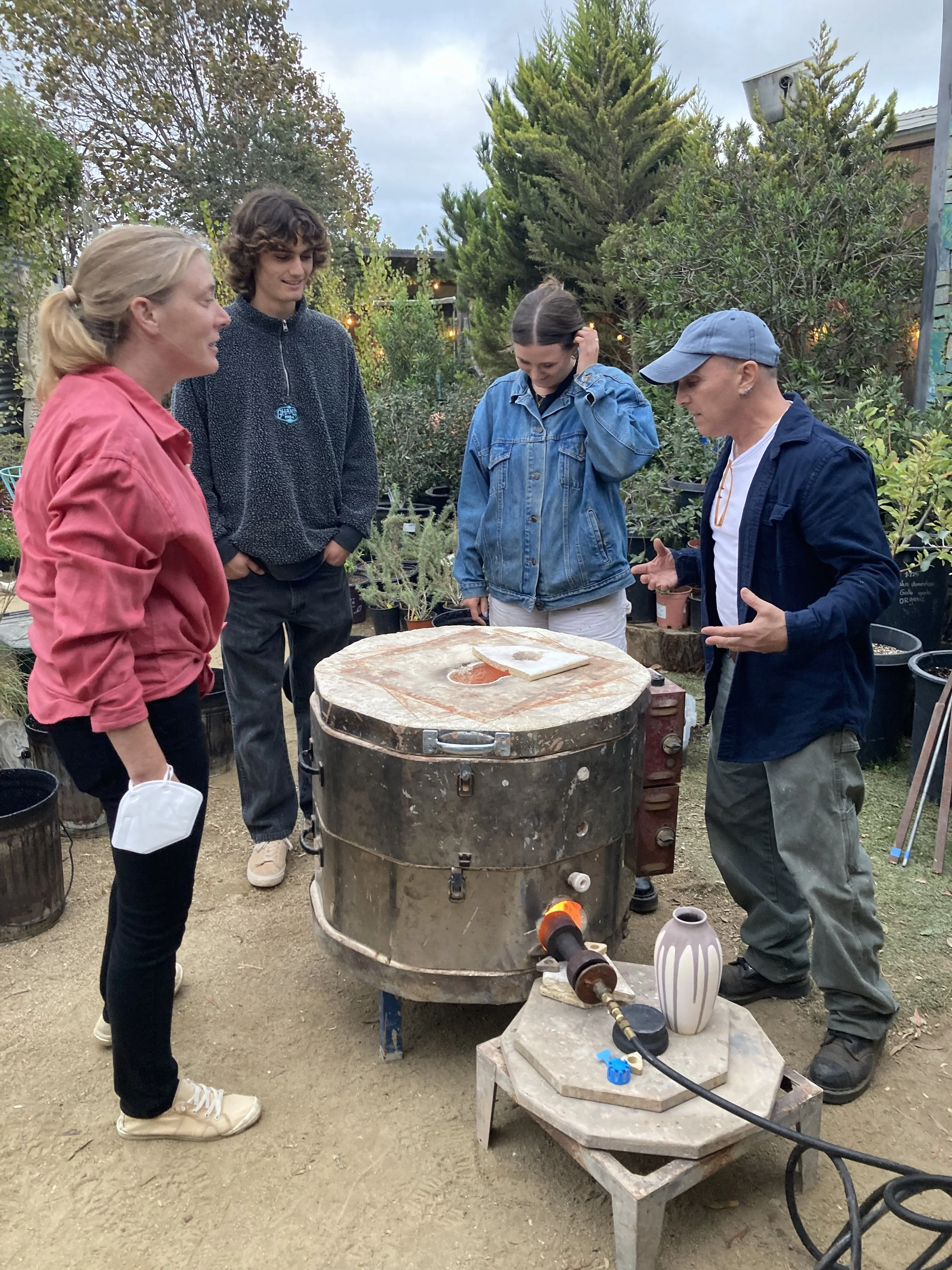 Four people standing around a pottery kiln outdoors, discussing pottery. The scene is in a garden with trees and plants in the background.