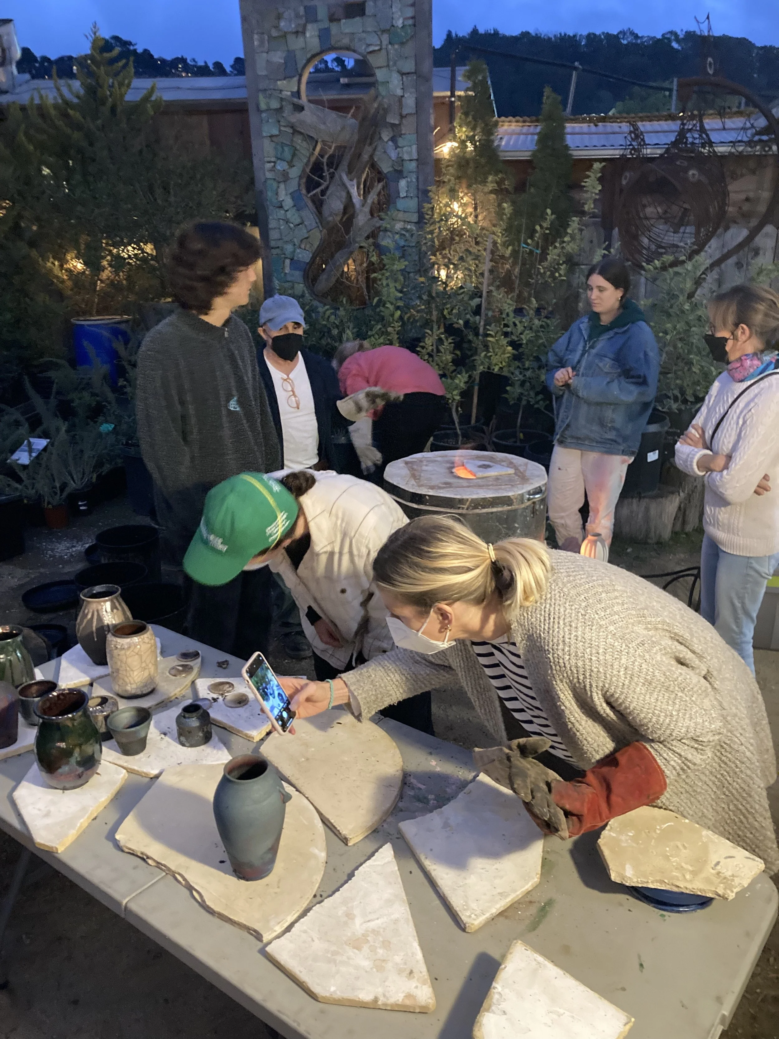 Raku workshop!  People outdoors at an evening pottery workshop, working on ceramic pieces on a long table, some taking photos, others examining the pottery, with plants and decorative art in the background.