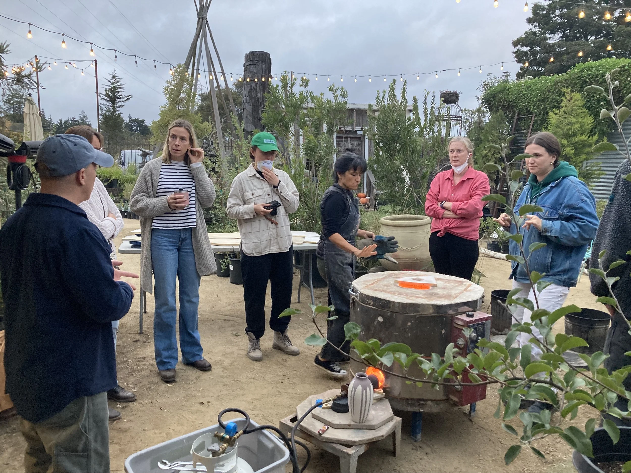 Group of people gathered outdoors around a pottery kiln at dusk, with string lights hanging overhead, some holding drinks, others with hands on their hips or taking notes, in a garden setting with greenery and trees in the background.