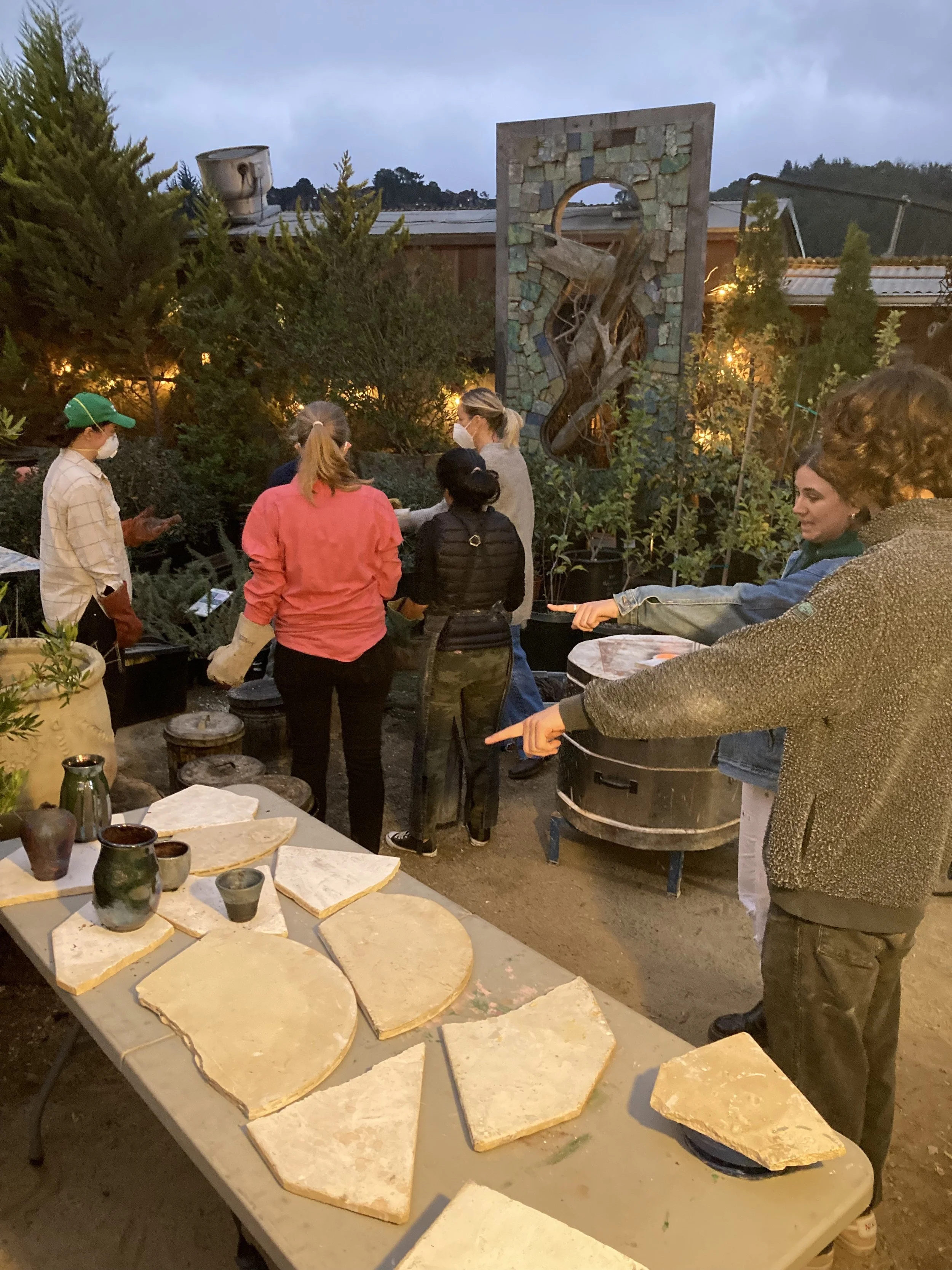 People gathered outdoors near a table with large stones, some cups, and plates, in a garden with trees and a decorative wall art installation in the background, during evening.