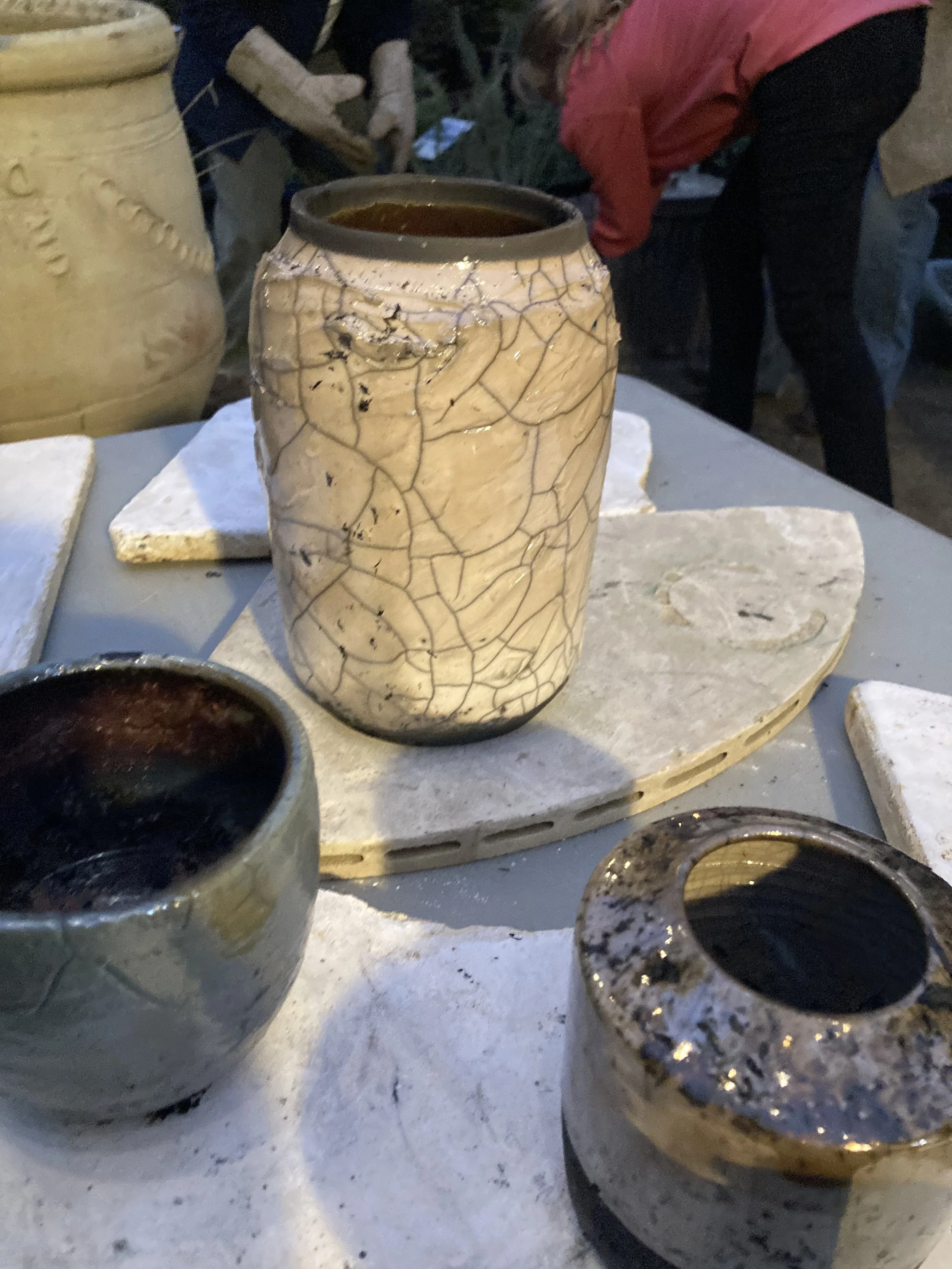A ceramic vase with a crackled glaze pattern, placed on a stone and surrounded by other pottery pieces and tiles.