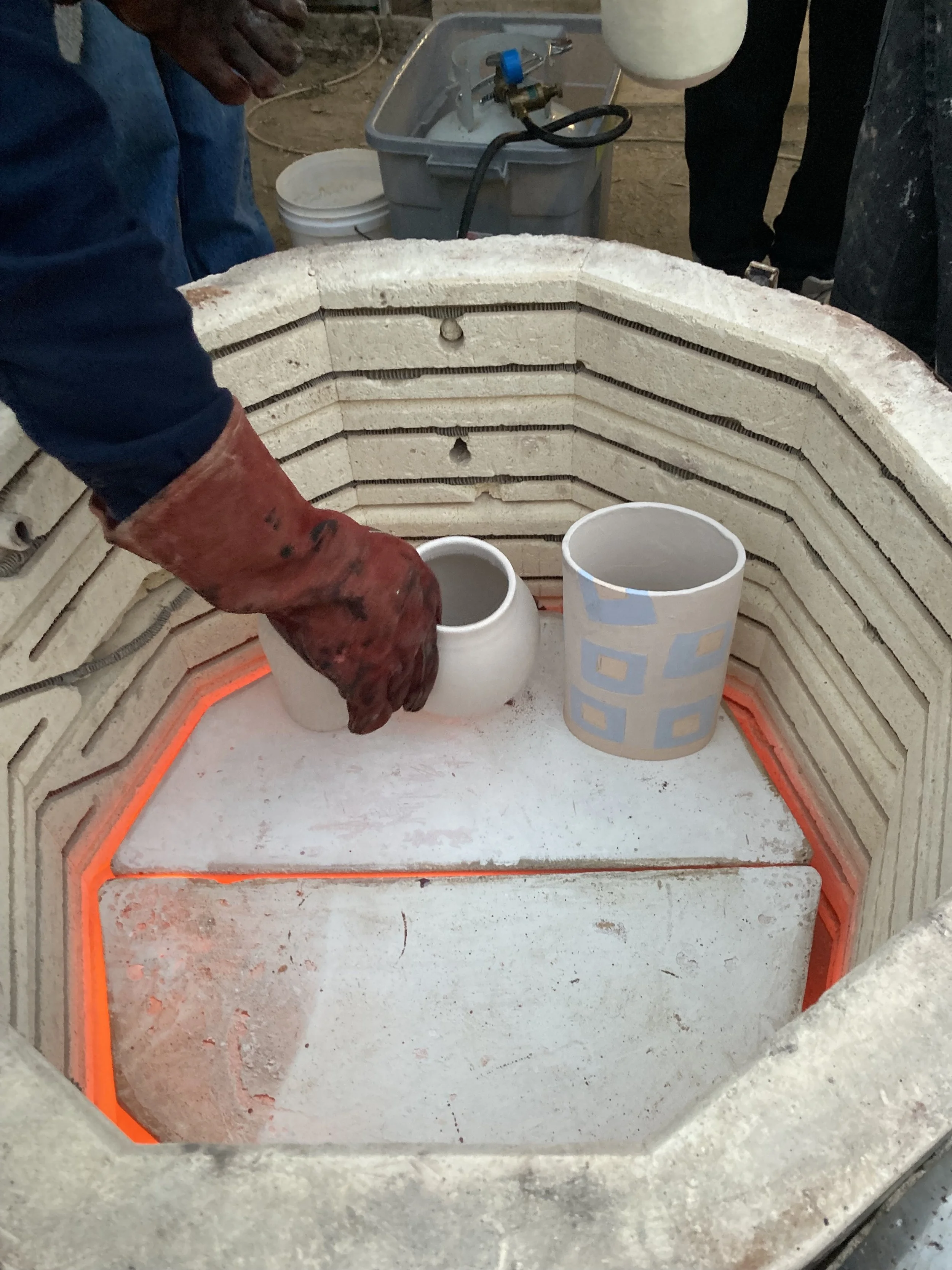 Person wearing red gloves handling a ceramic piece inside a kiln with glowing orange heating elements. The kiln's interior is lined with refractory bricks and has two ceramic pieces, one plain and one painted, being fused or fired.