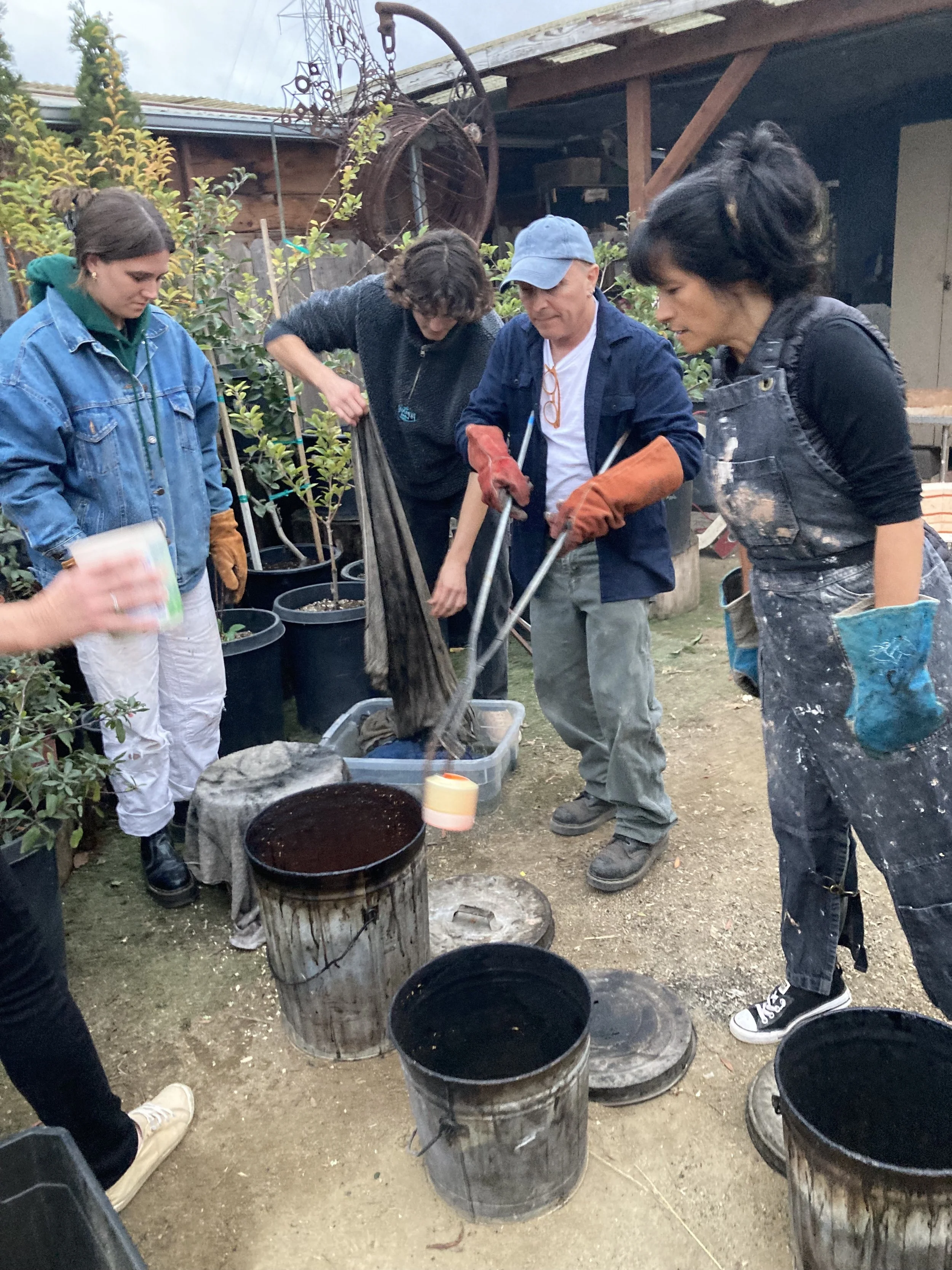 A group of five people gathered outdoors in a garden, involved in a woodworking or craft project using heat or dye, with buckets and tools around them.