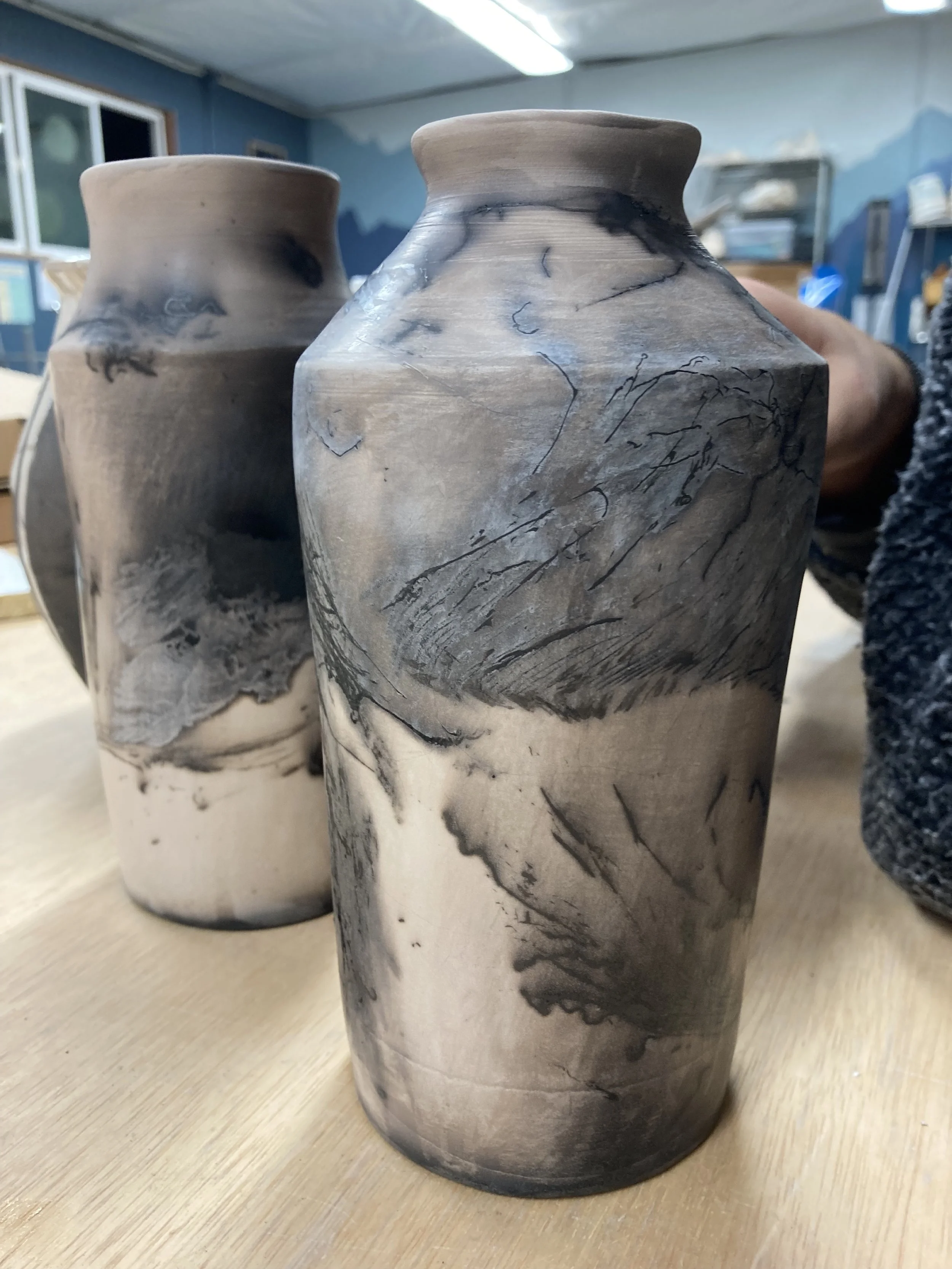 Two raku ceramic vases with marbled black, gray, and cream patterns placed on a wooden surface, inside a pottery studio.