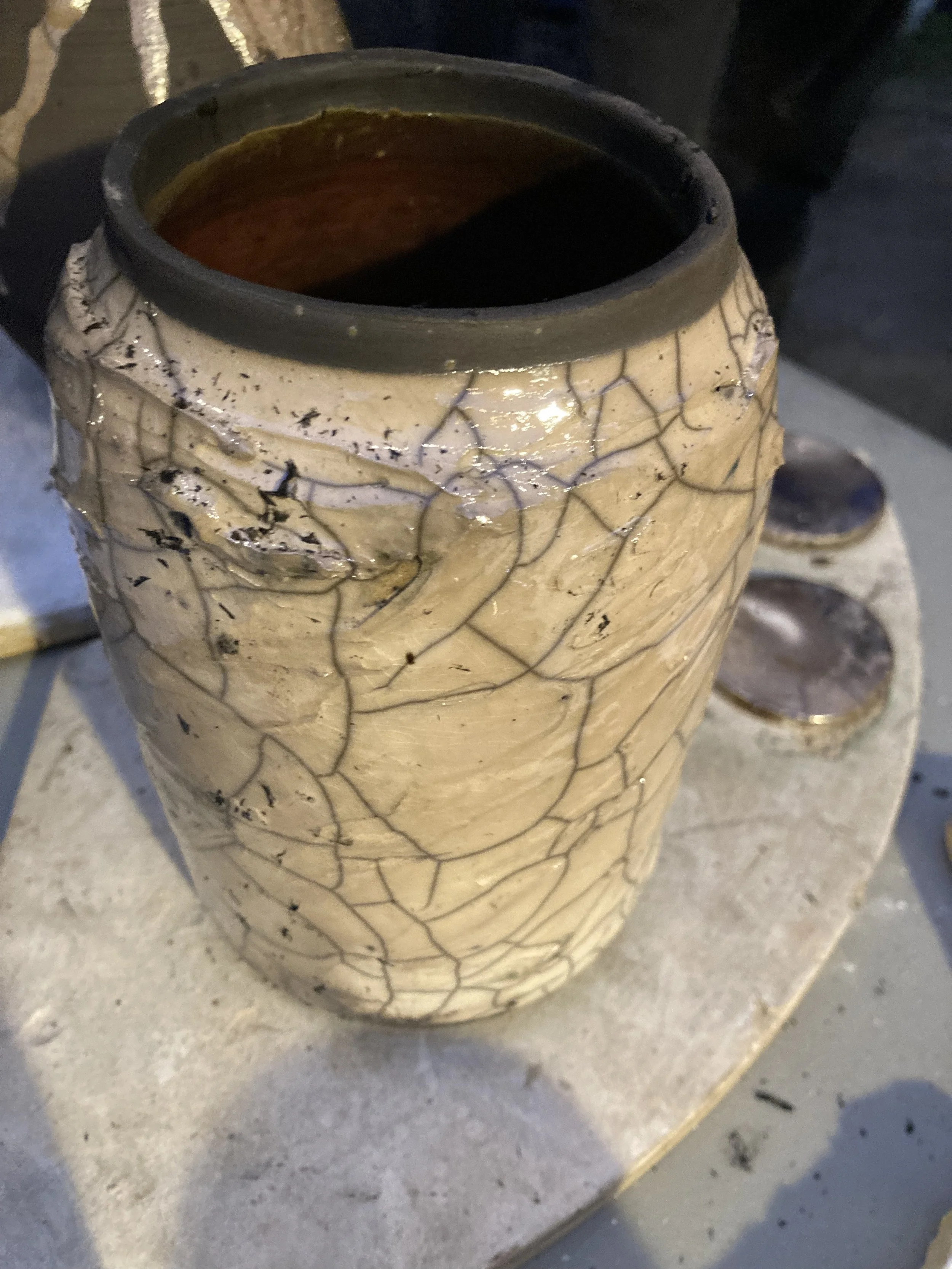 A tall, beige ceramic vase with a crackled glaze design, placed on a stone or concrete surface. The vase's opening shows a dark interior.