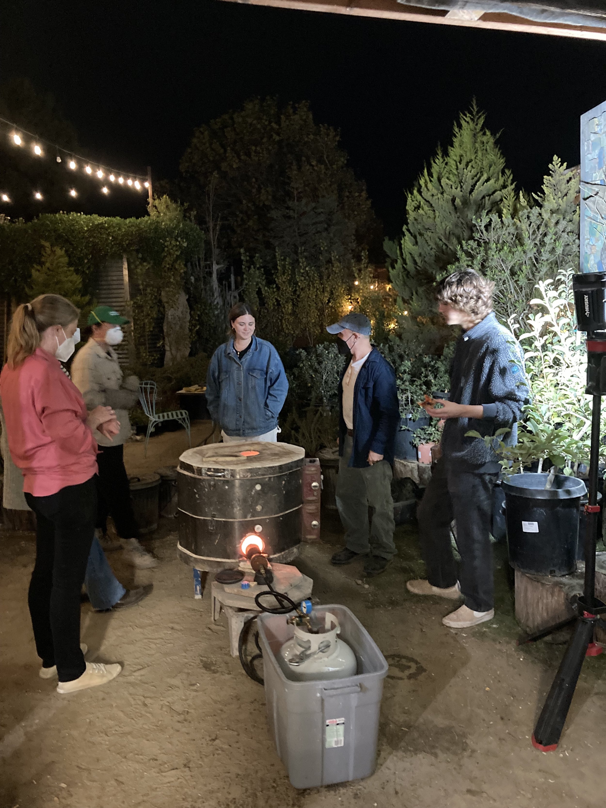 Group of five people gathered around a forge outdoors at night, some wearing masks, with trees and string lights in the background.