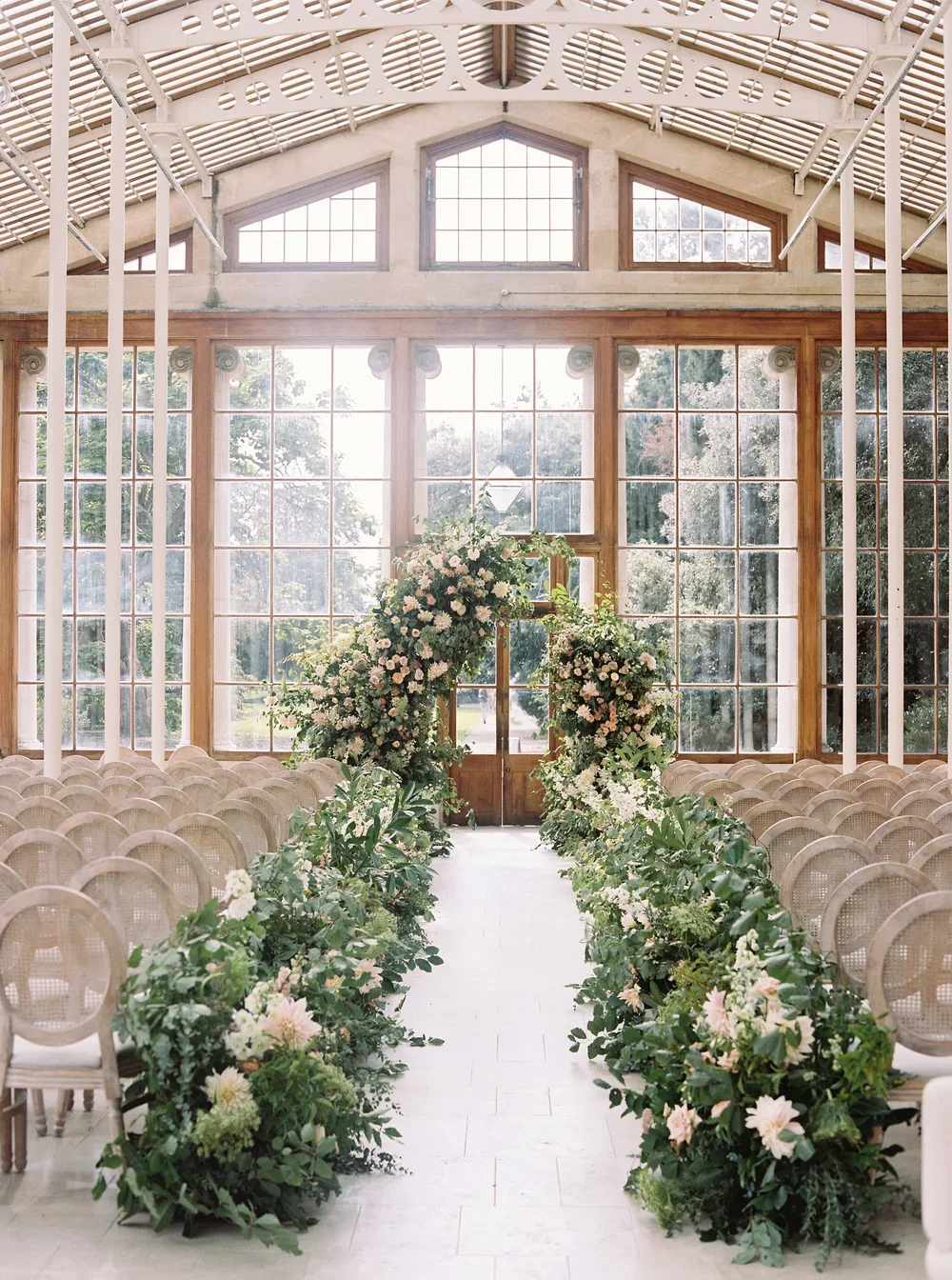 Bright indoor wedding ceremony space with large windows, decorated floral aisle, and archway with pink and white flowers.