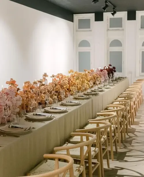 Luxury brand event dining setup featuring natural wood wishbone chairs, long tables and peach floral arrangements.