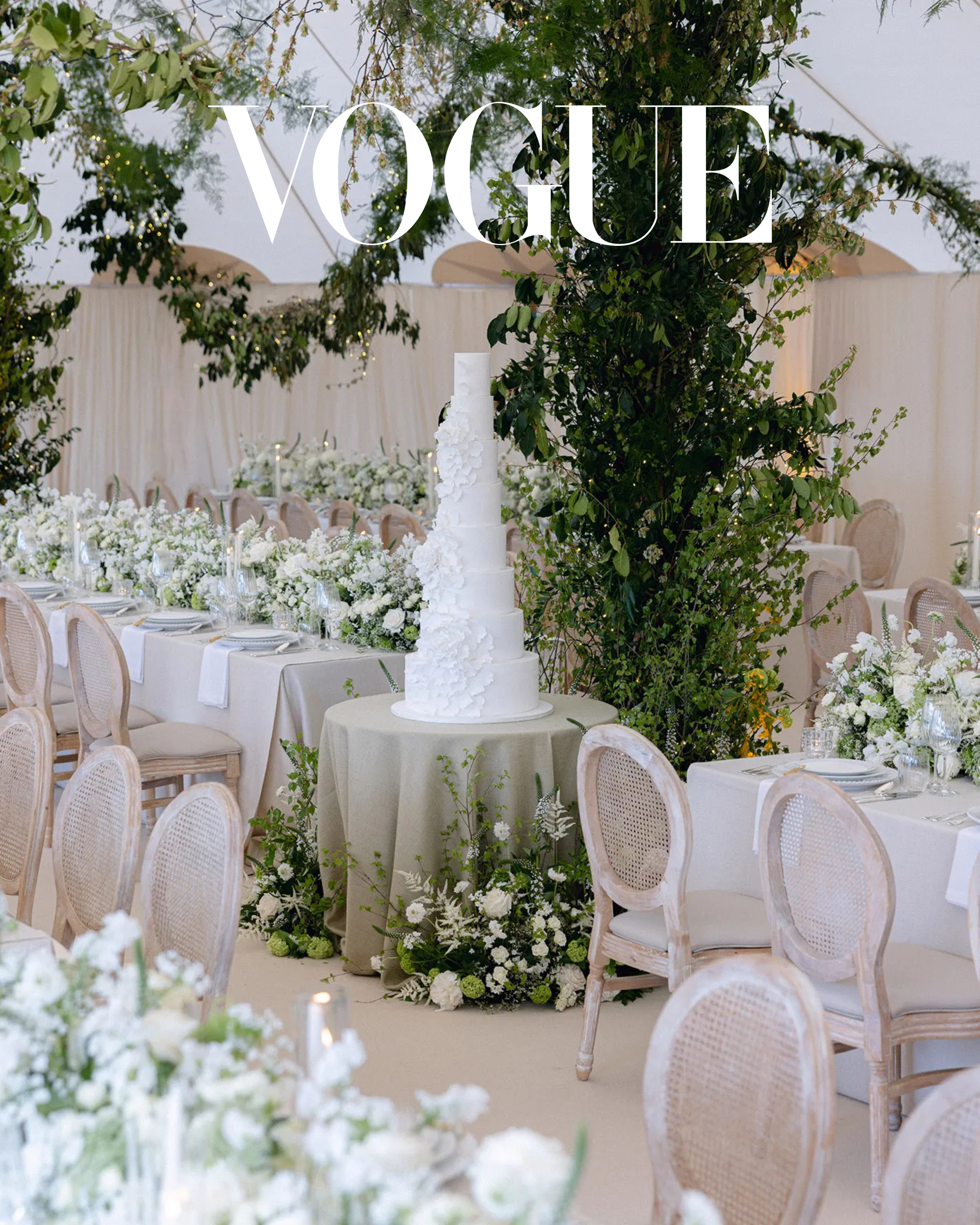 Space within the marquee filled with rows of upholstered caneback chairs, foraged foliage installations, soft neutral draping and a large white wedding cake