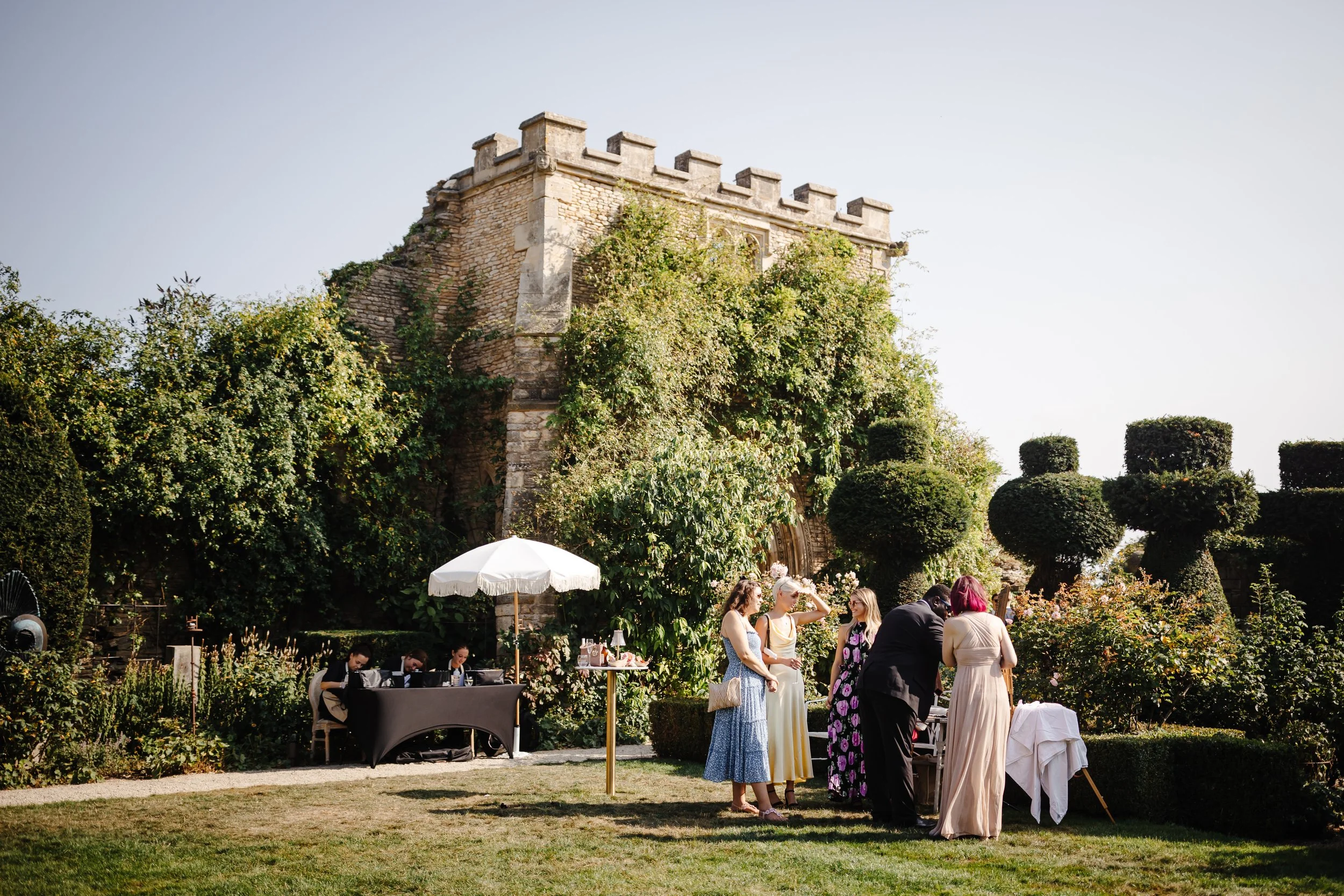 People dressed in formal attire at an outdoor gathering or celebration in front of a historic stone structure, with a table, umbrella, and well-manicured bushes and trees.