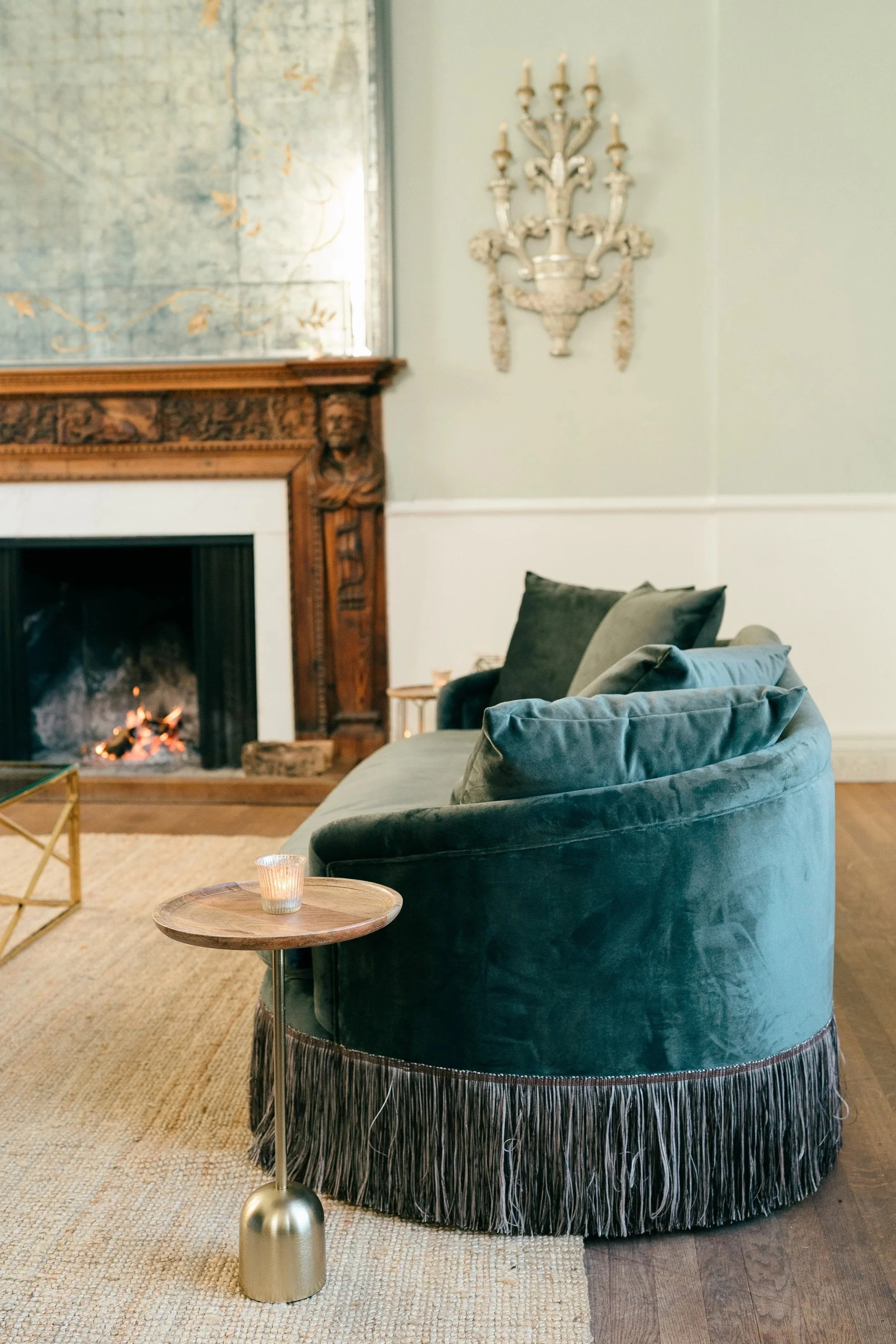 A cozy living room with a green velvet tufted sofa, a small wooden side table with a candle, a fireplace with a carved wooden mantle, a painting above the fireplace, and decorative wall sconces.