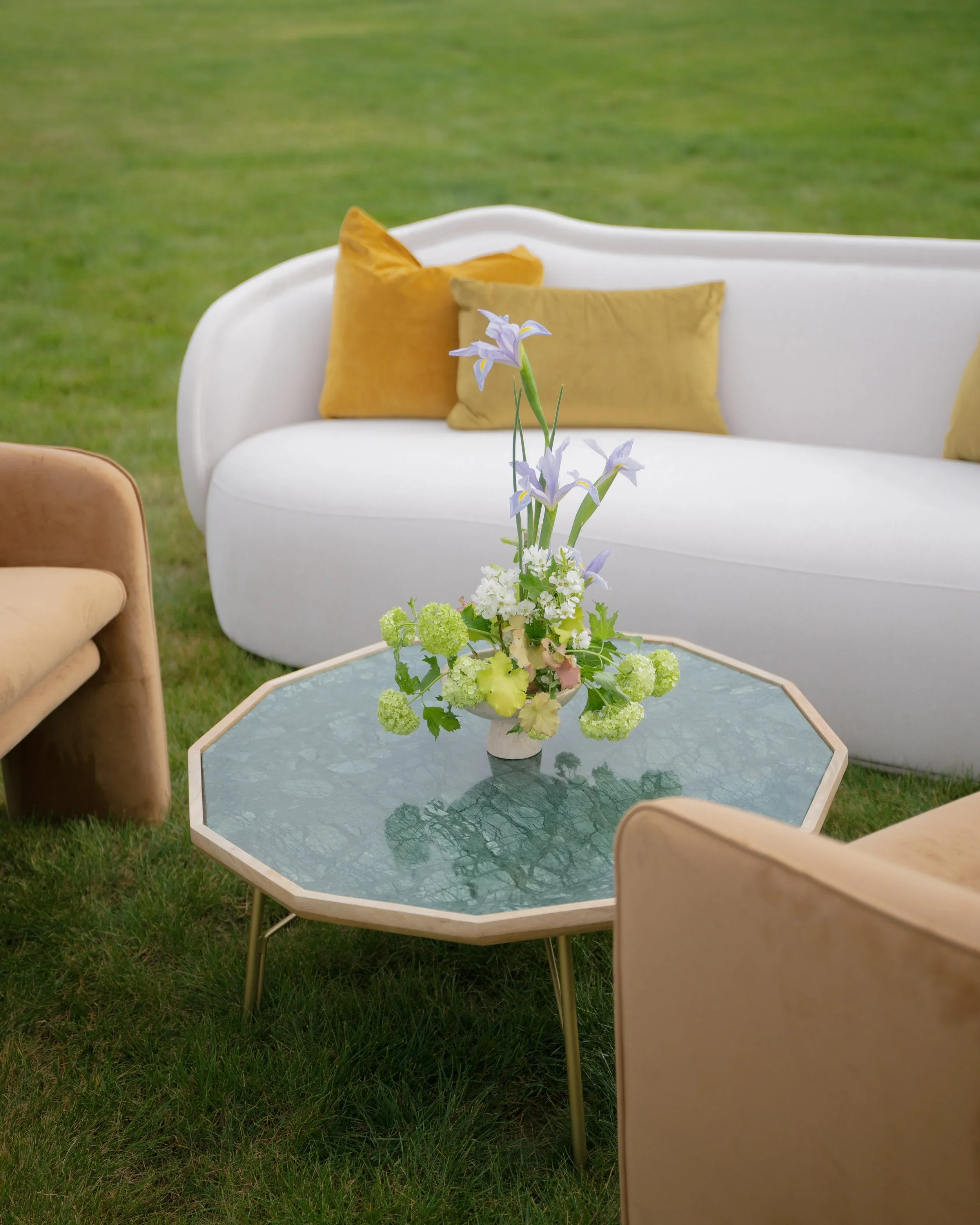 An outdoor seating area with a white curved sofa with yellow cushions, a glass-topped coffee table, and a floral arrangement with purple irises and white and green flowers on the table, set on green grass.