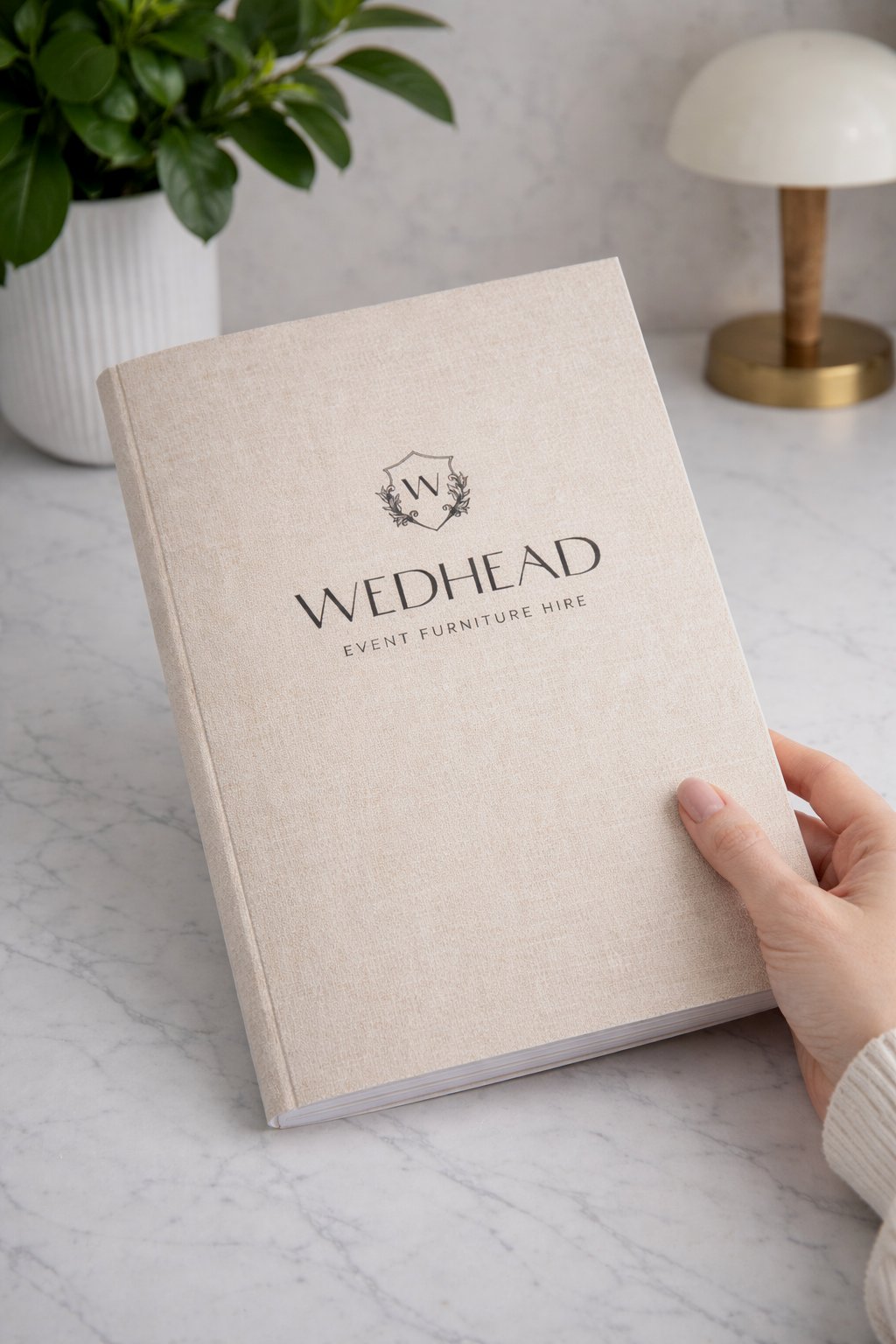 A person holding a beige book titled 'WEDHEAD Event Furniture Hire' on a marble tabletop, with a white ribbed plant pot containing green foliage and a modern white and brass table lamp in the background.