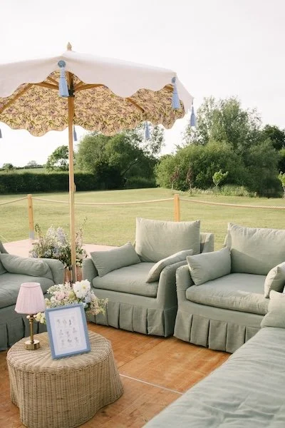 Elegant wedding outdoor lounge area with green farmhouse slipcover famrhouse sofas, parasols and a rattan outdoor coffee table with a wireless lamp on top