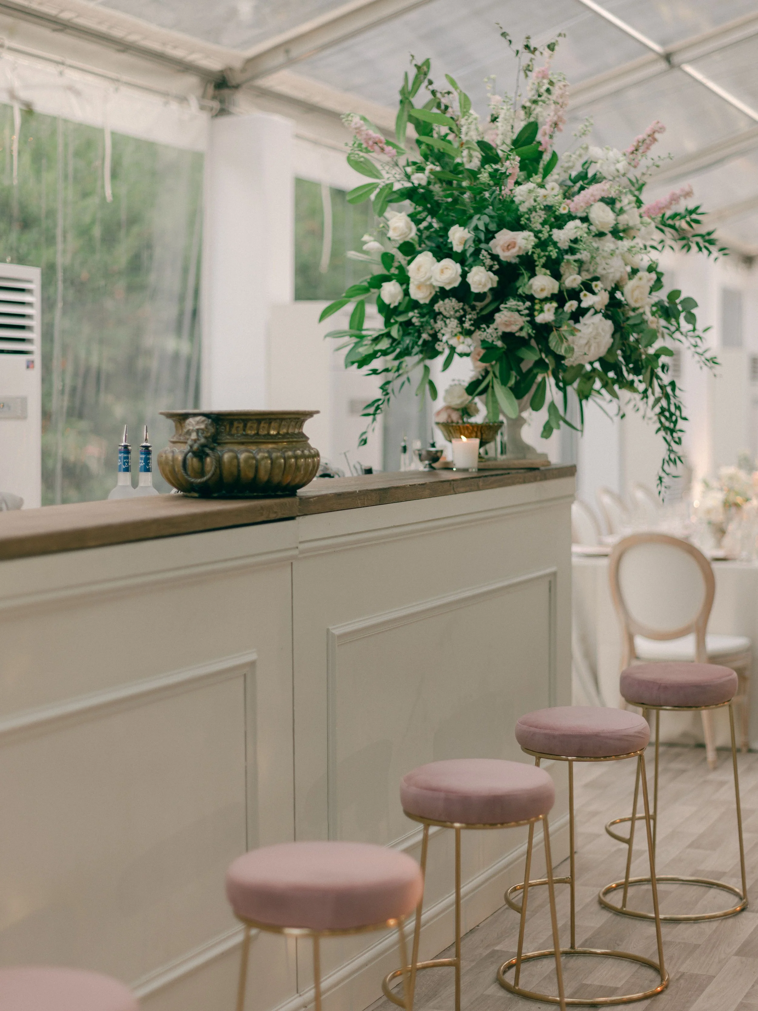 Elegant event decoration featuring a large floral arrangement of white roses and pink flowers on a bar counter with pink velvet stools and a candle.
