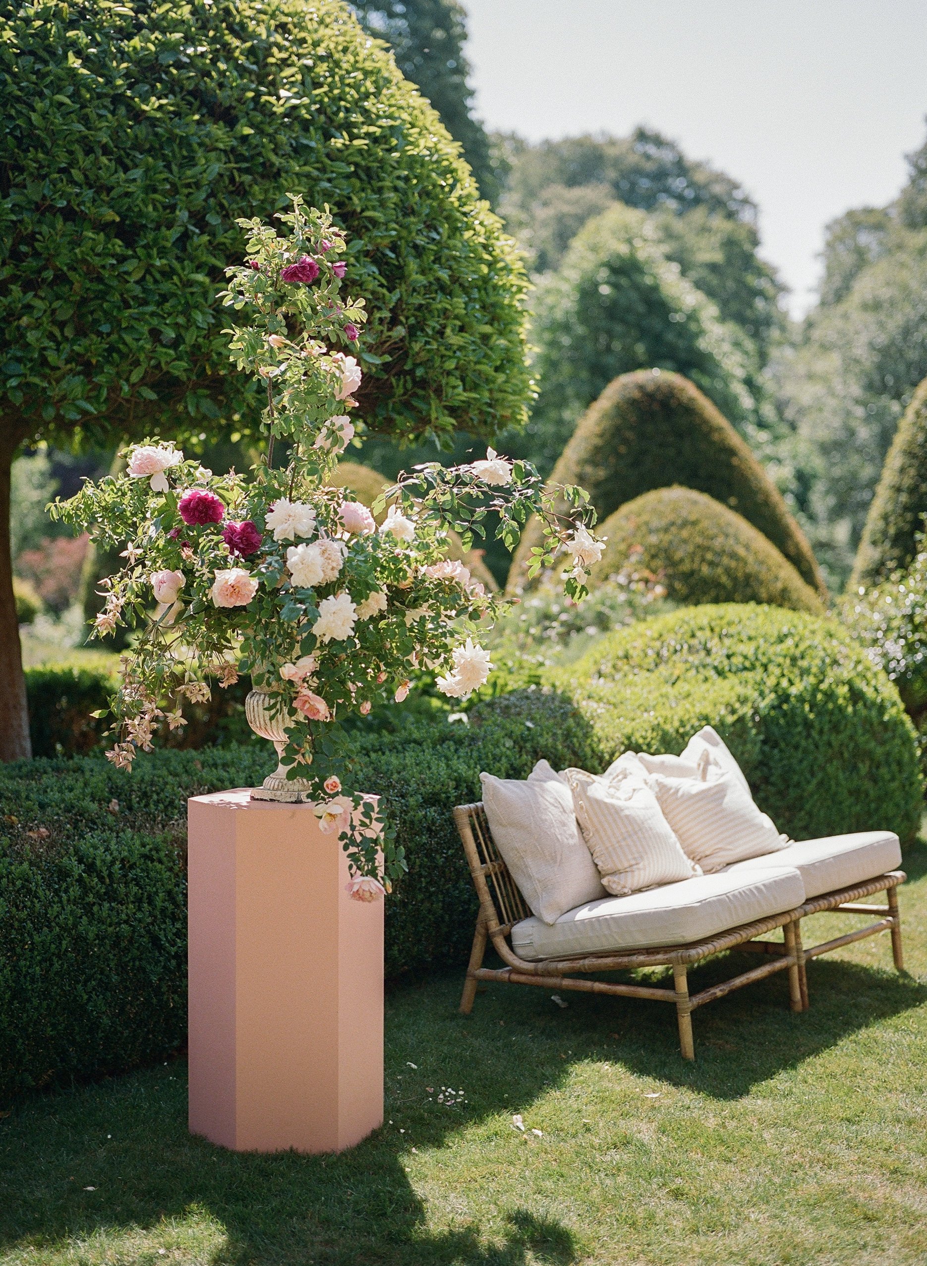 A garden scene with a wicker bench with white cushions, a pink pedestal with a large floral arrangement, neatly trimmed bushes, and trees in the background, under a bright sky.