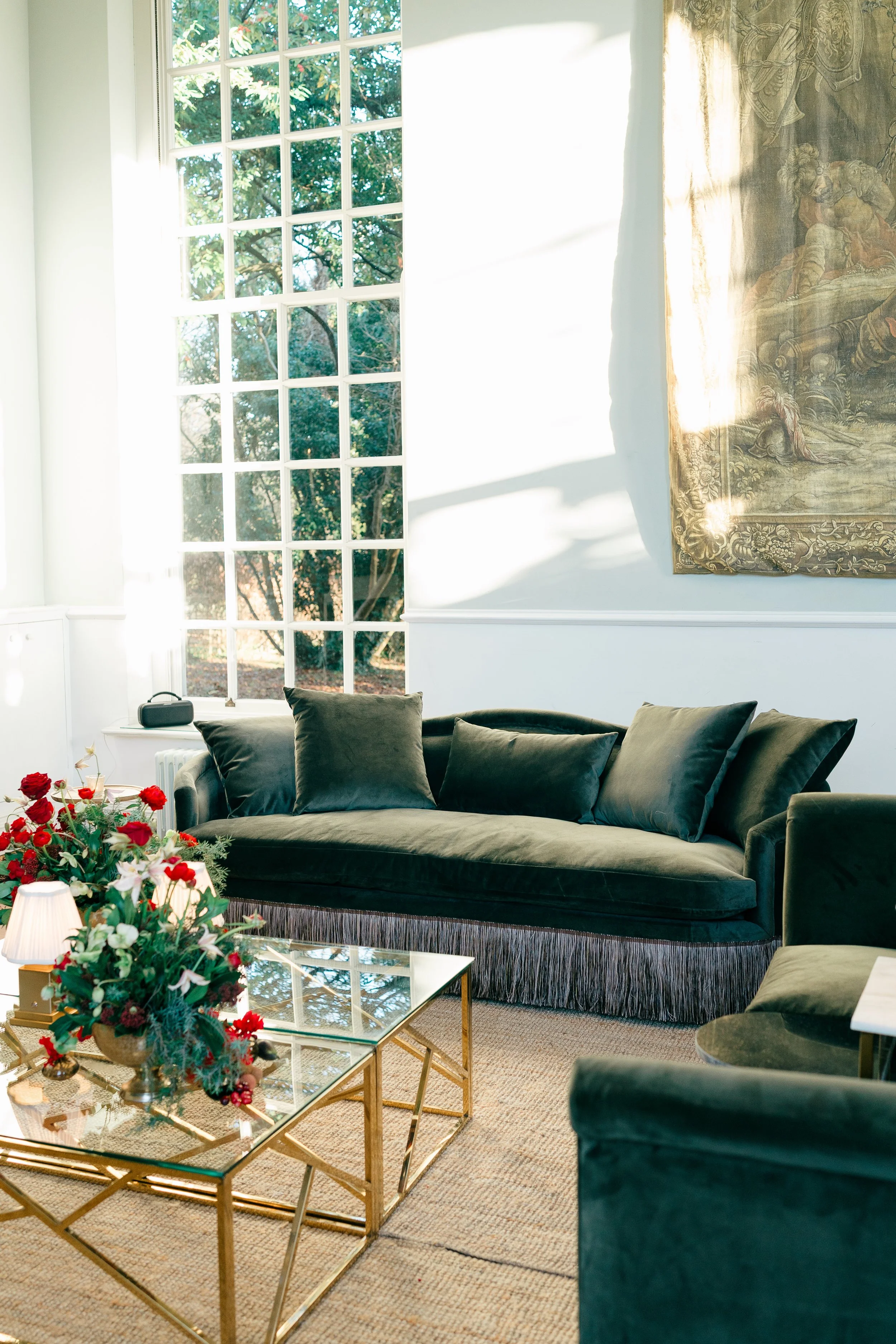 Sunlit living room with a large window, a dark green velvet sofa with matching cushions, a glass coffee table with a floral arrangement, and a tapestry hanging on the wall.