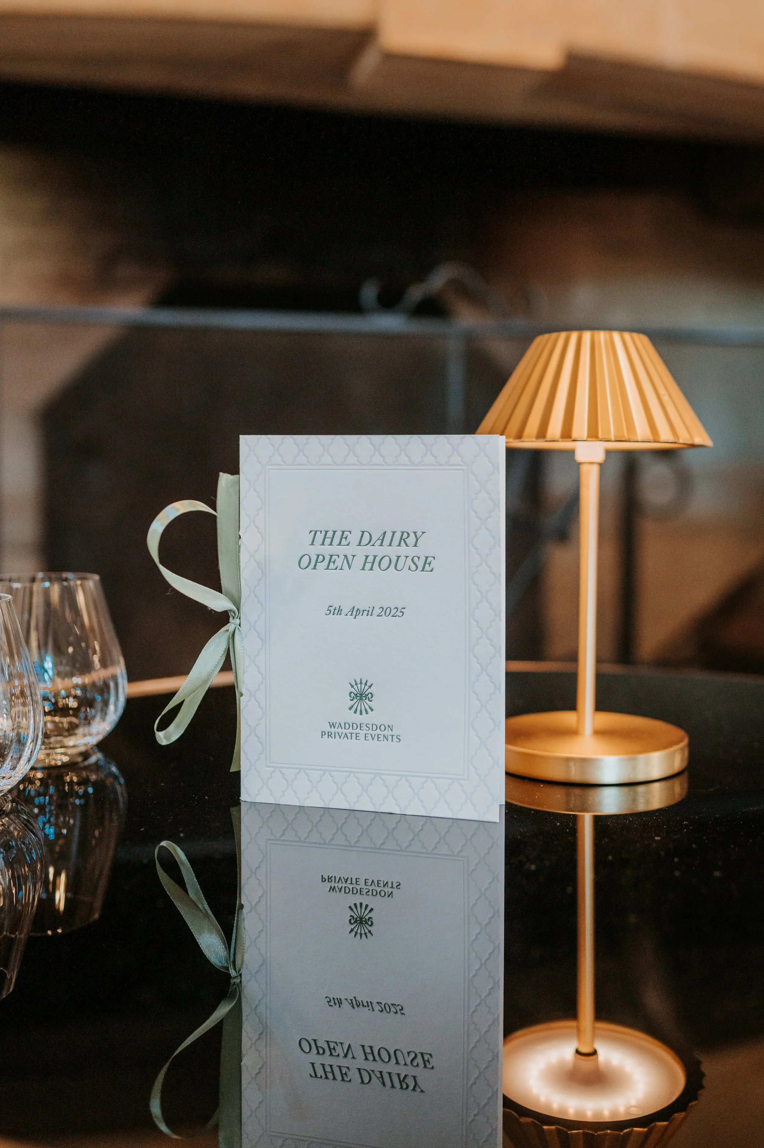 A table with a white program that reads 'The Dairy Open House, 5th April 2025, Waddesdon Private Events,' a gold-colored lamp, and several empty wine glasses.
